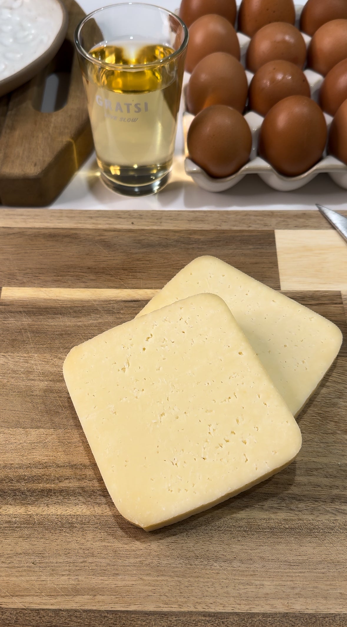 Two slices of pale yellow cheese are placed on a wooden cutting board, with brown eggs in a carton and a glass of white wine in the background.