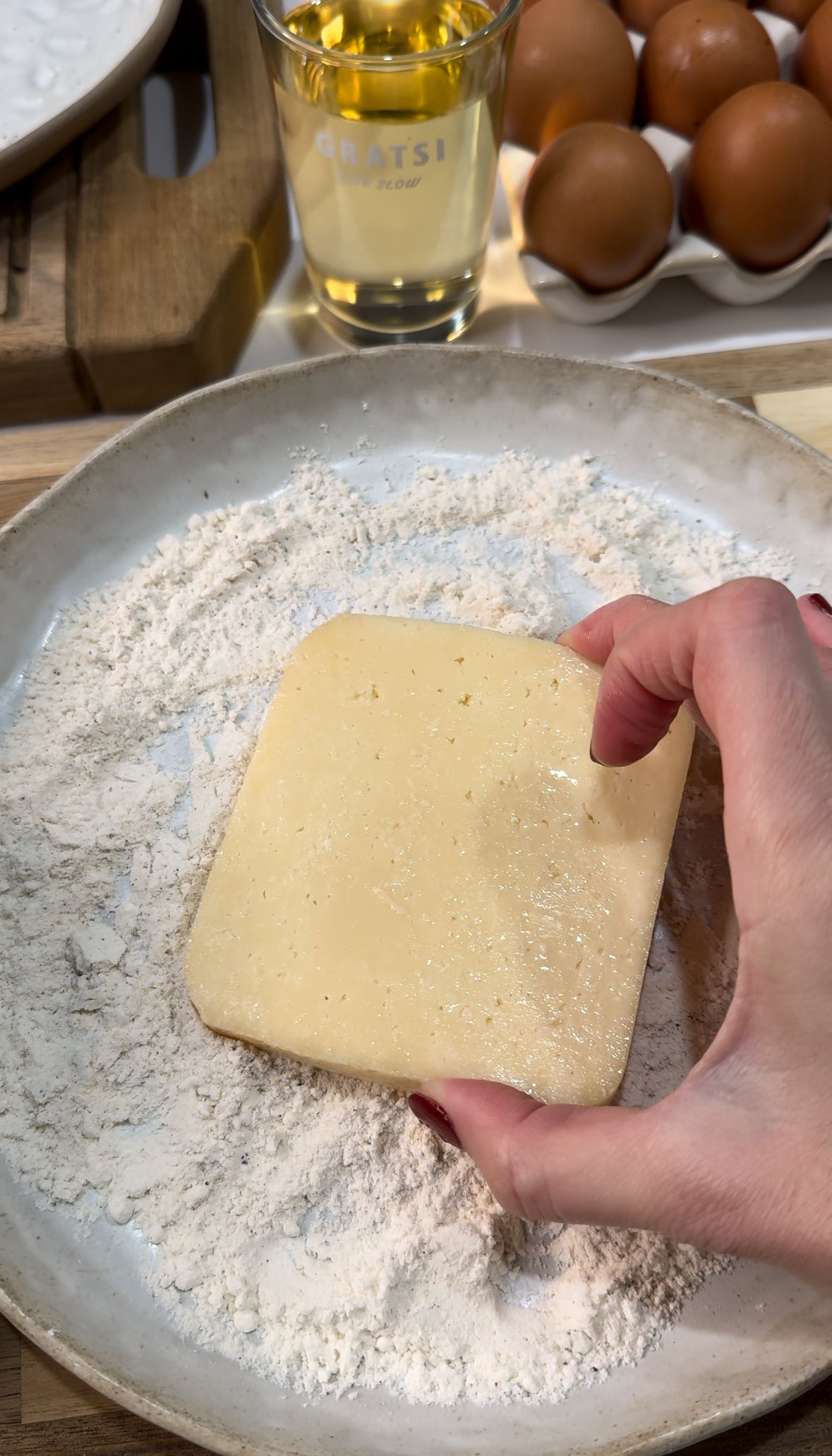 A hand dips a rectangular slice of cheese into a plate of flour. Nearby are eggs, a glass of liquid, and a wooden cutting board.