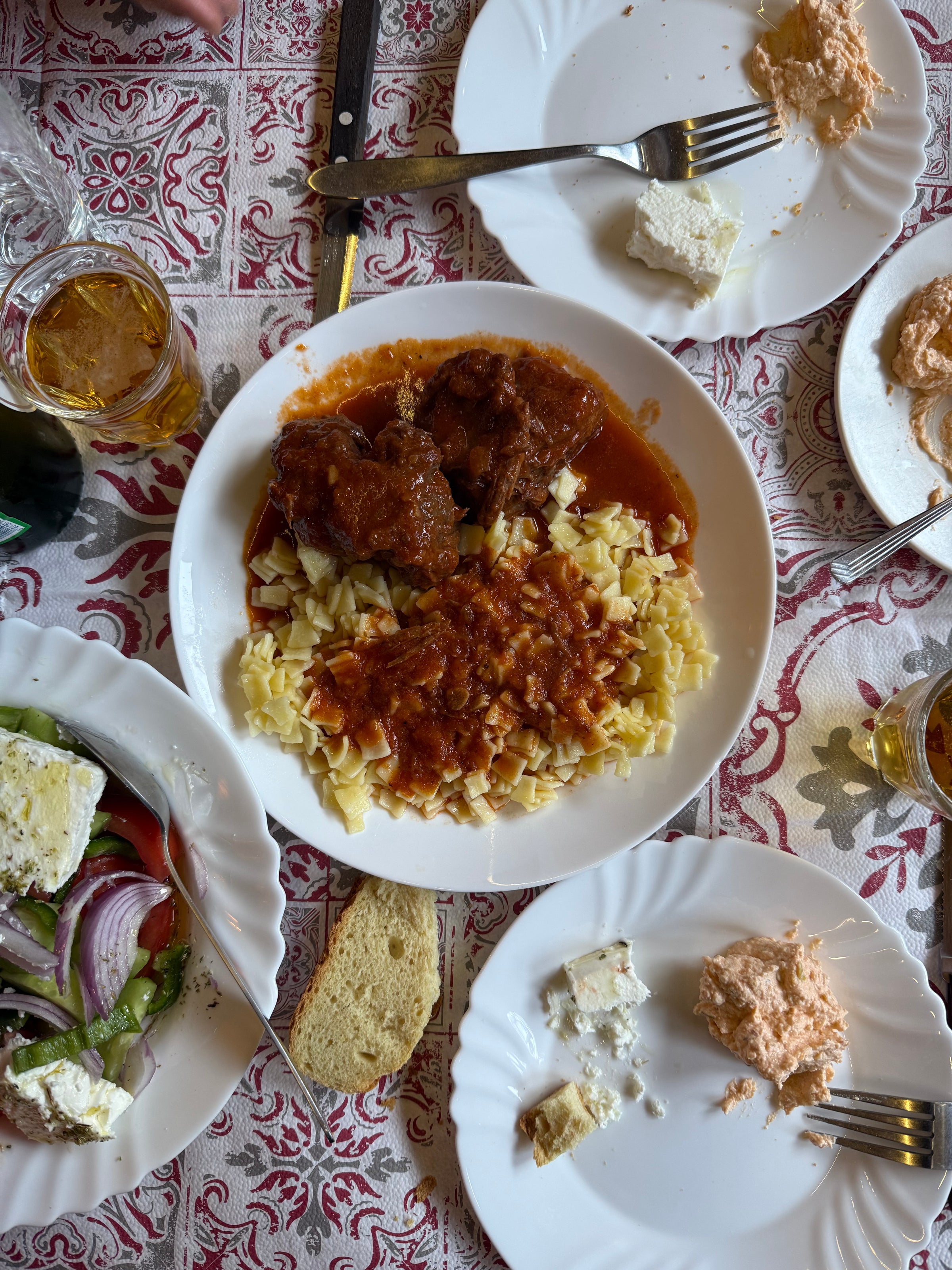 A table set with a main dish of pasta and meat in tomato sauce, plates with cheese spreads, bread slices, a Greek salad, and drinks on a patterned red and white tablecloth.