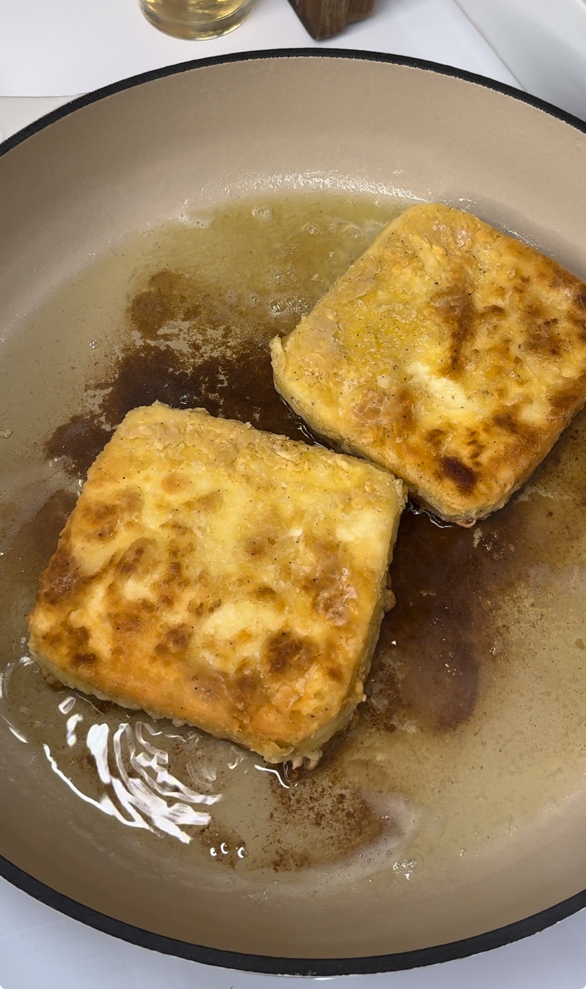 Two golden-brown breaded squares are being fried in oil in a beige skillet, with the oil bubbling around them.