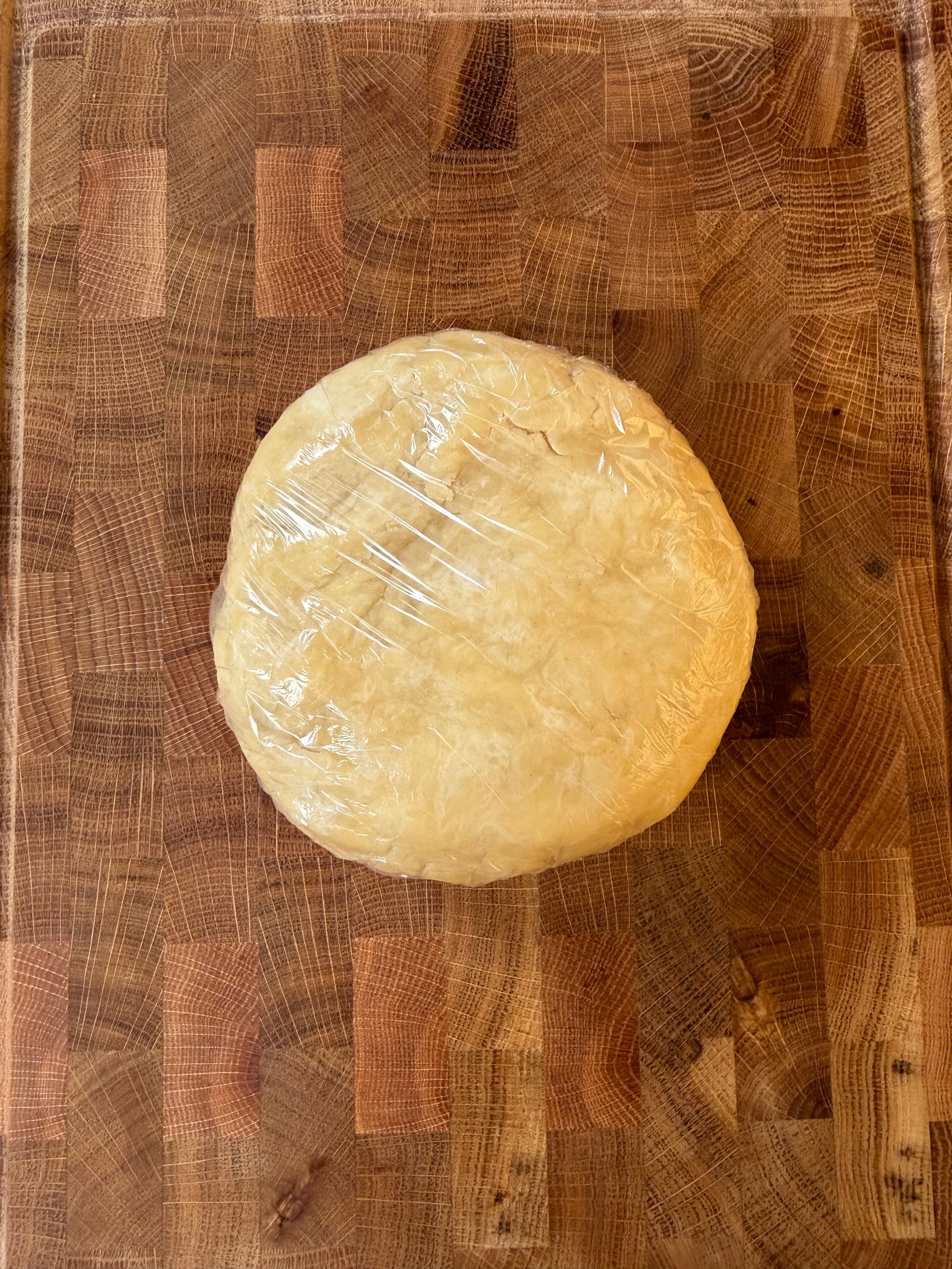 A disk of dough wrapped in plastic on a wooden cutting board.