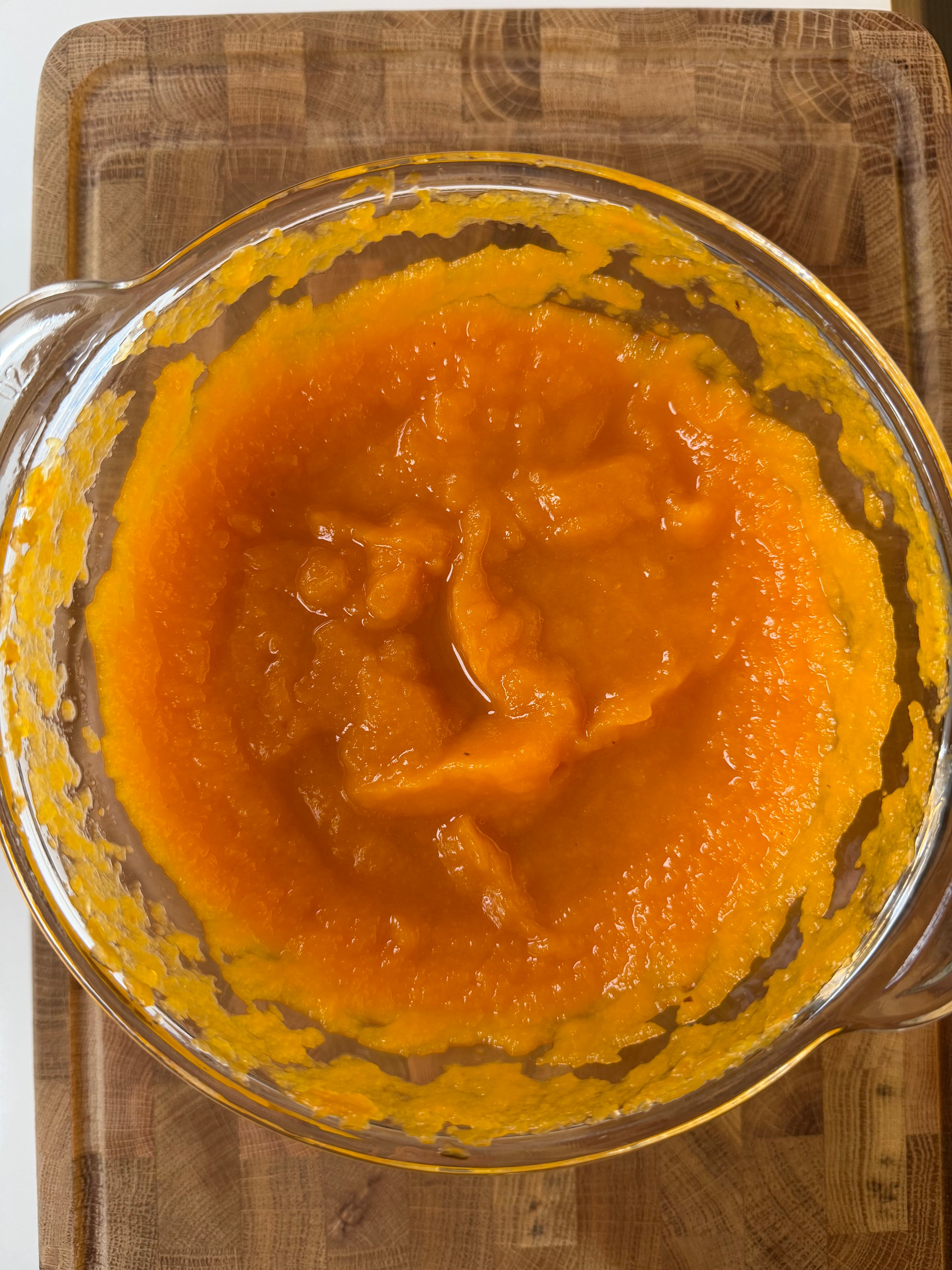A glass bowl filled with orange fruit or vegetable purée sitting on a wooden cutting board.
