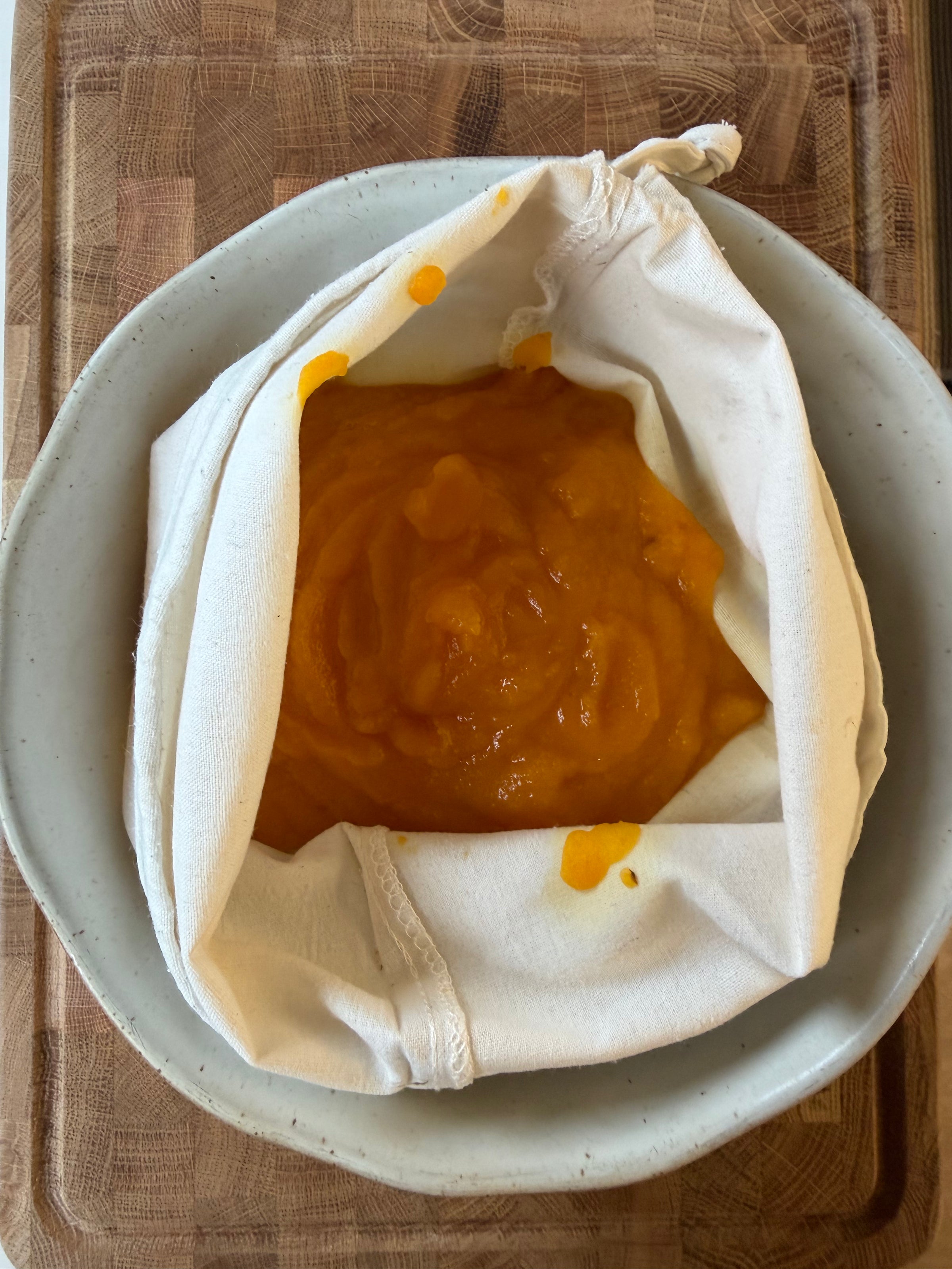 A light-colored fabric nut milk bag containing bright orange pureed squash inside a shallow bowl.