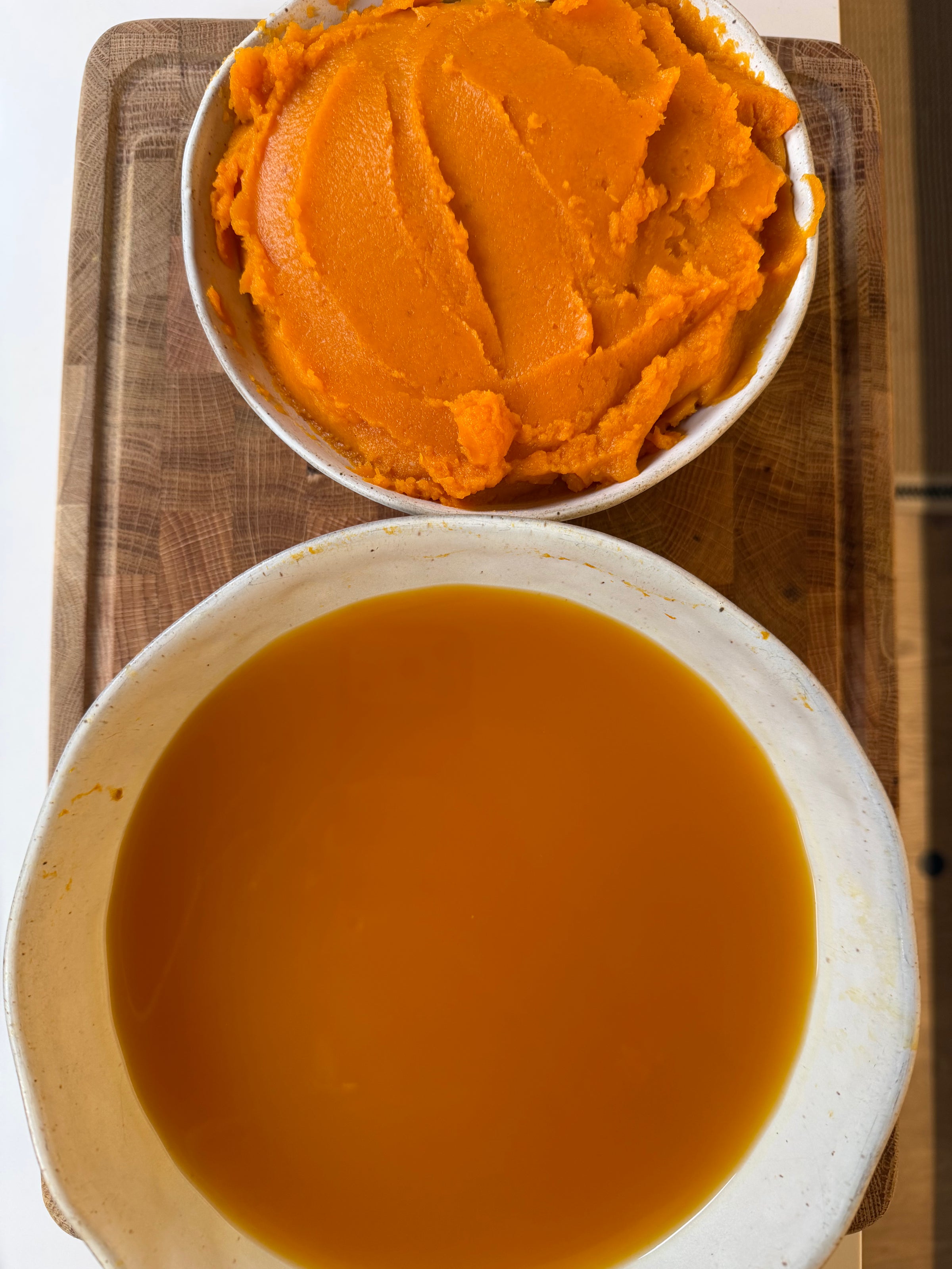 Two bowls on a wooden cutting board: one filled with orange pumpkin puree and the other with orange soup.