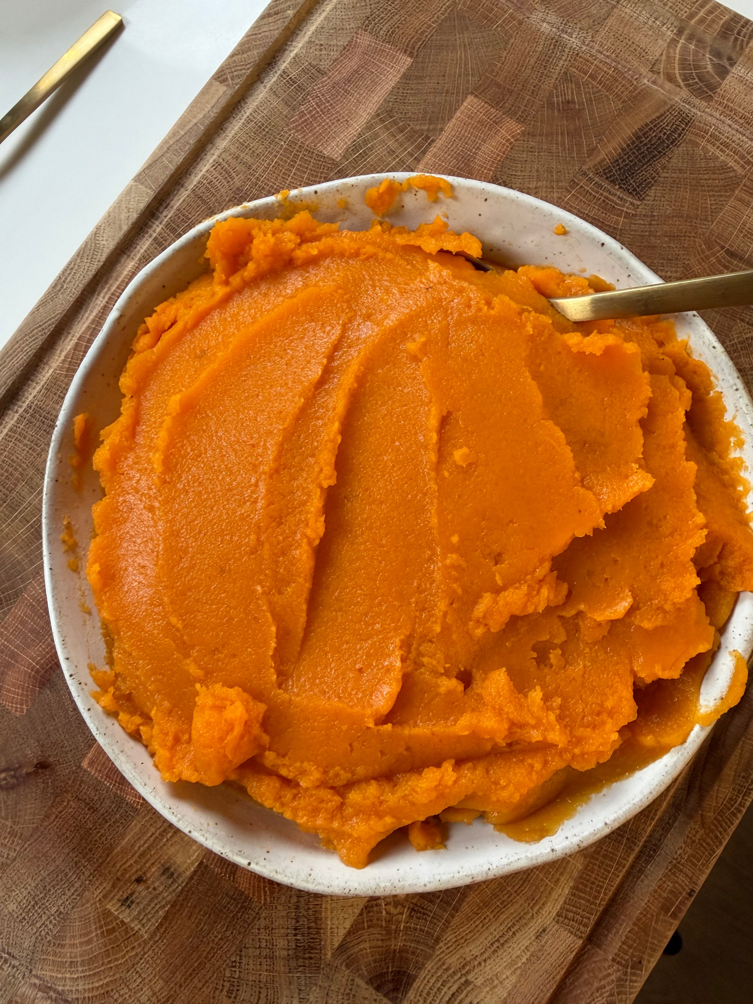 A bowl of mashed sweet potatoes on a wooden cutting board with a gold spoon.