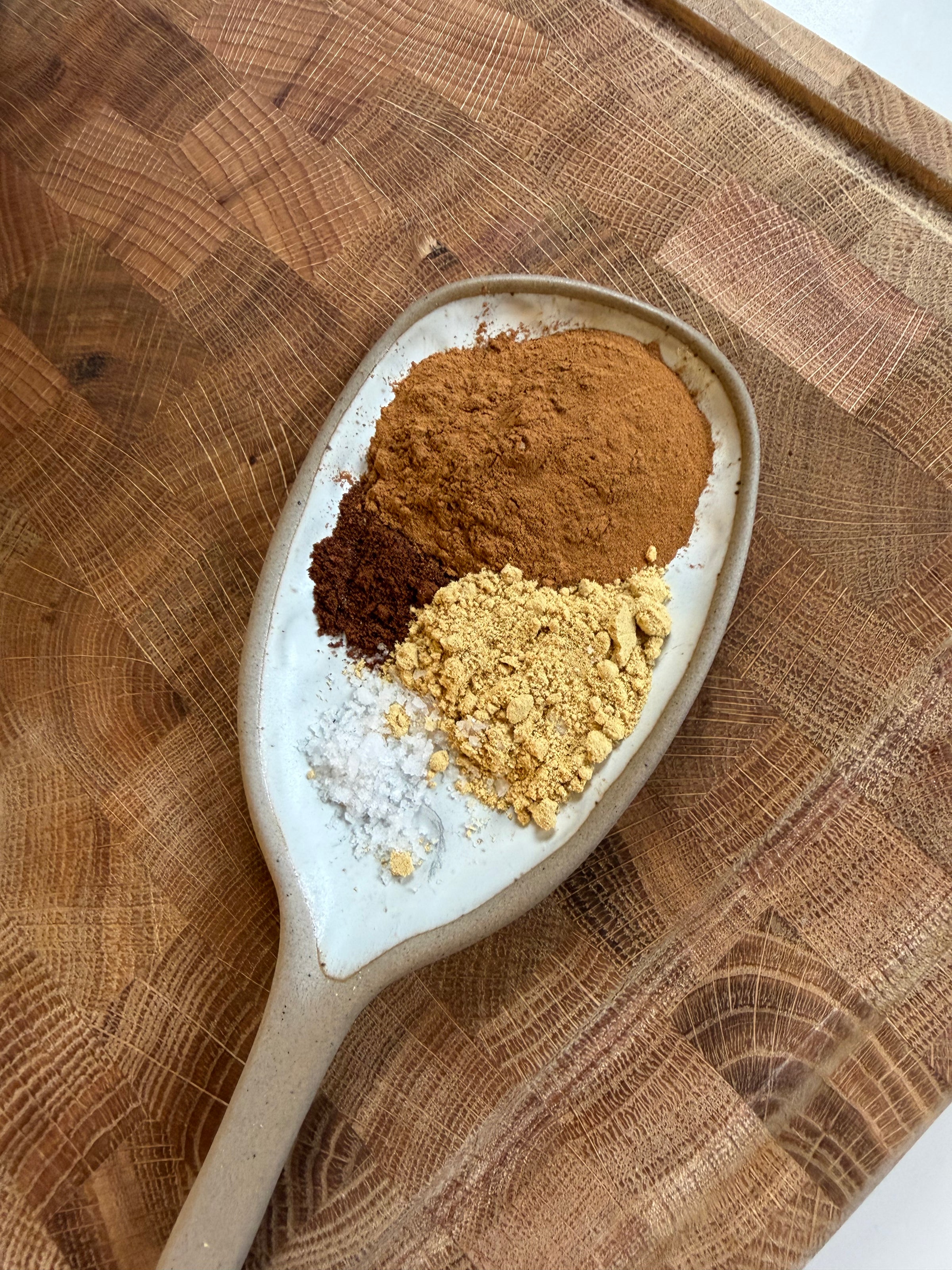 A ceramic spoon on a wooden board holding small mounds of cinnamon, cocoa powder, ground ginger, and coarse salt.