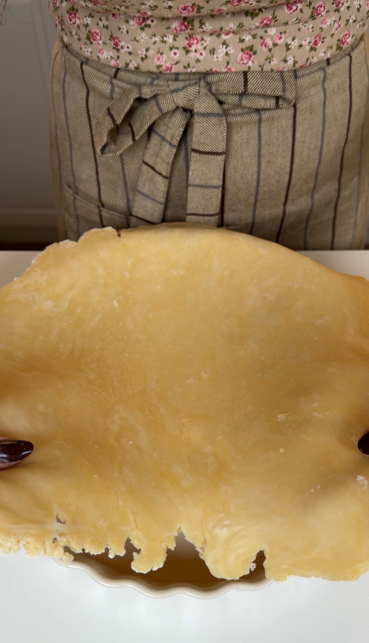 A person wearing a striped apron holds a sheet of raw pie dough over a baking dish.