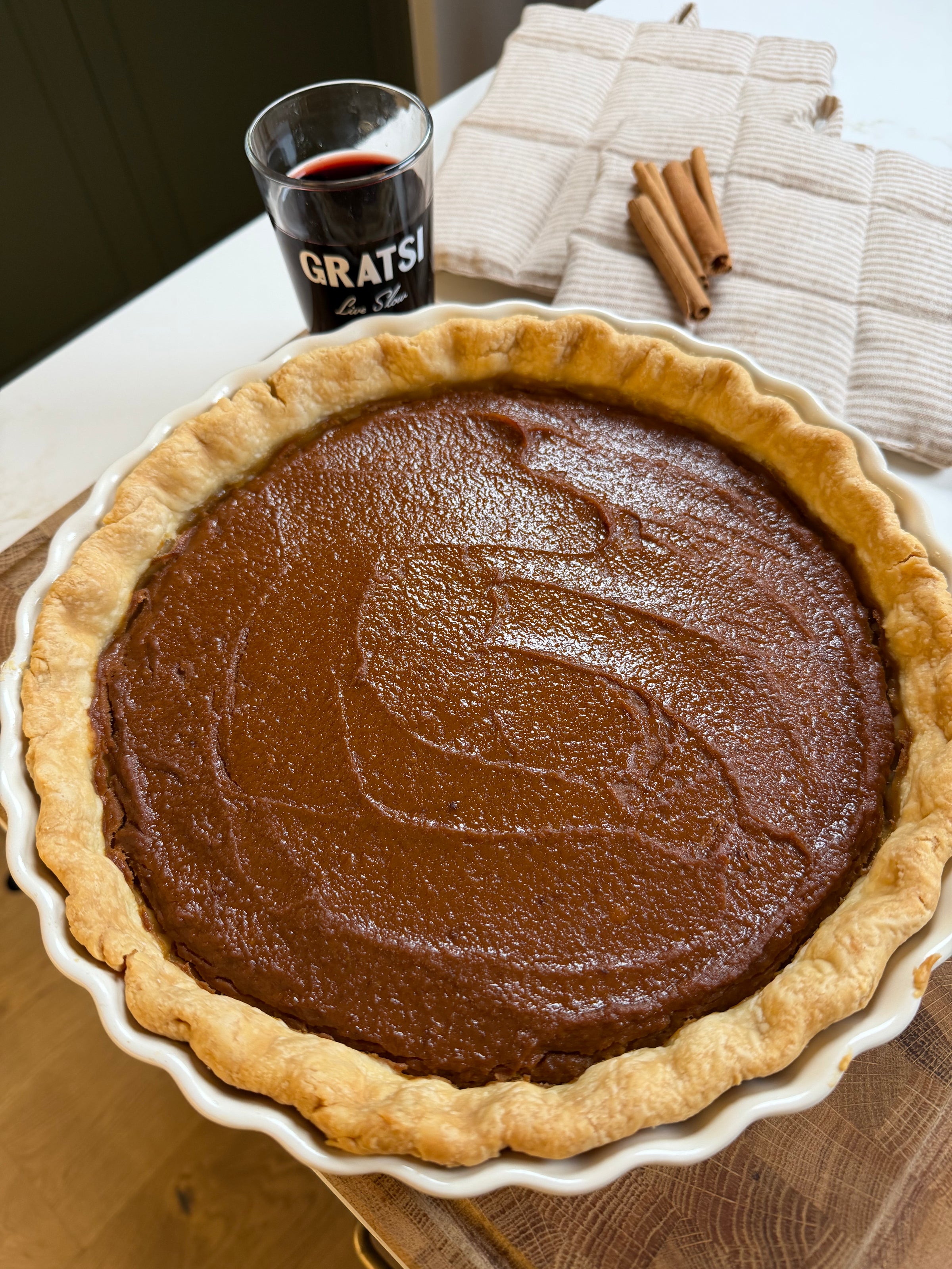 A baked pumpkin pie in a scalloped white ceramic dish, with a glass of red wine and two cinnamon sticks nearby.