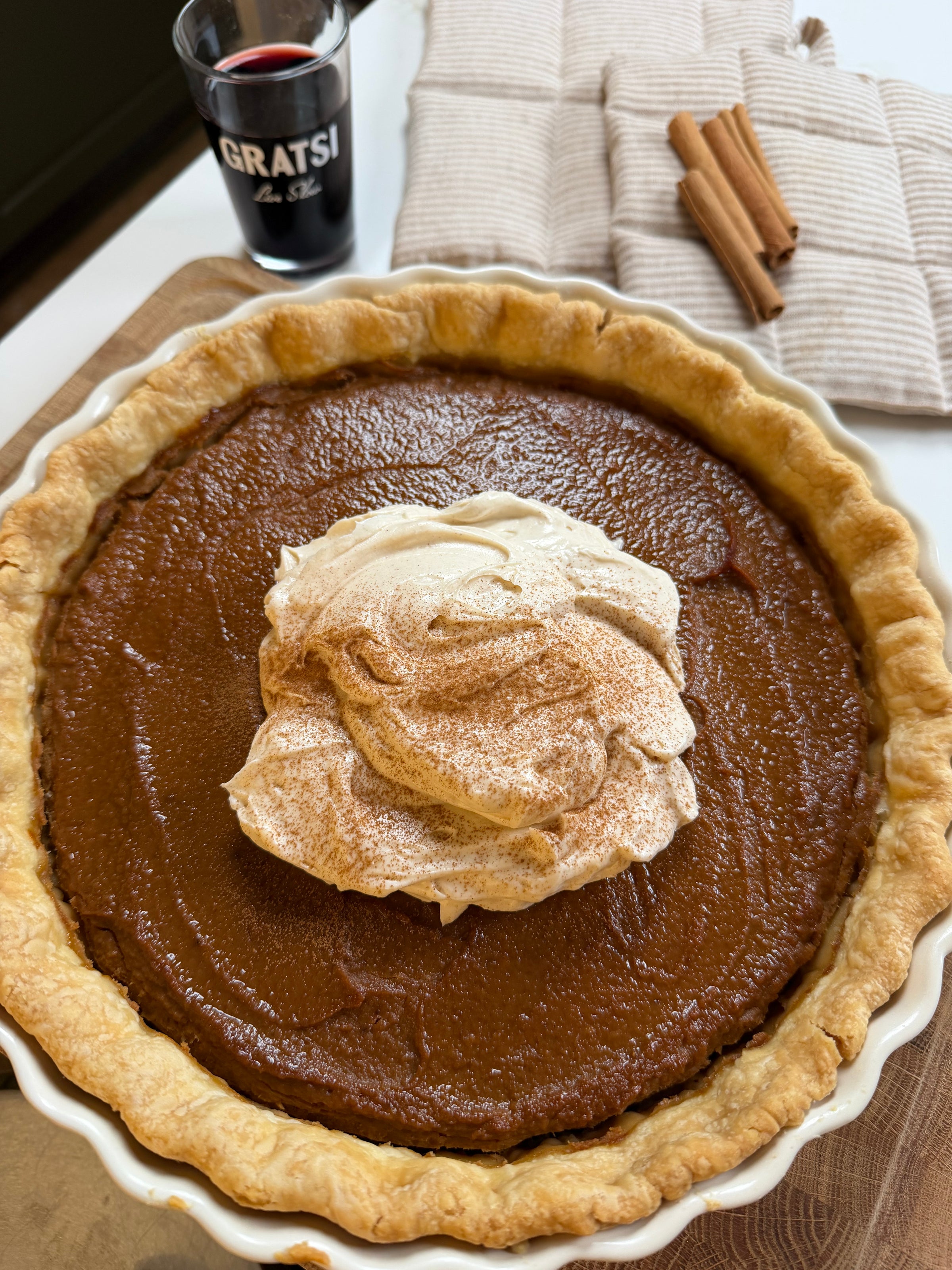 Pumpkin pie in a ceramic dish topped with a dollop of whipped cream and cinnamon, with a glass of red wine and cinnamon sticks nearby.