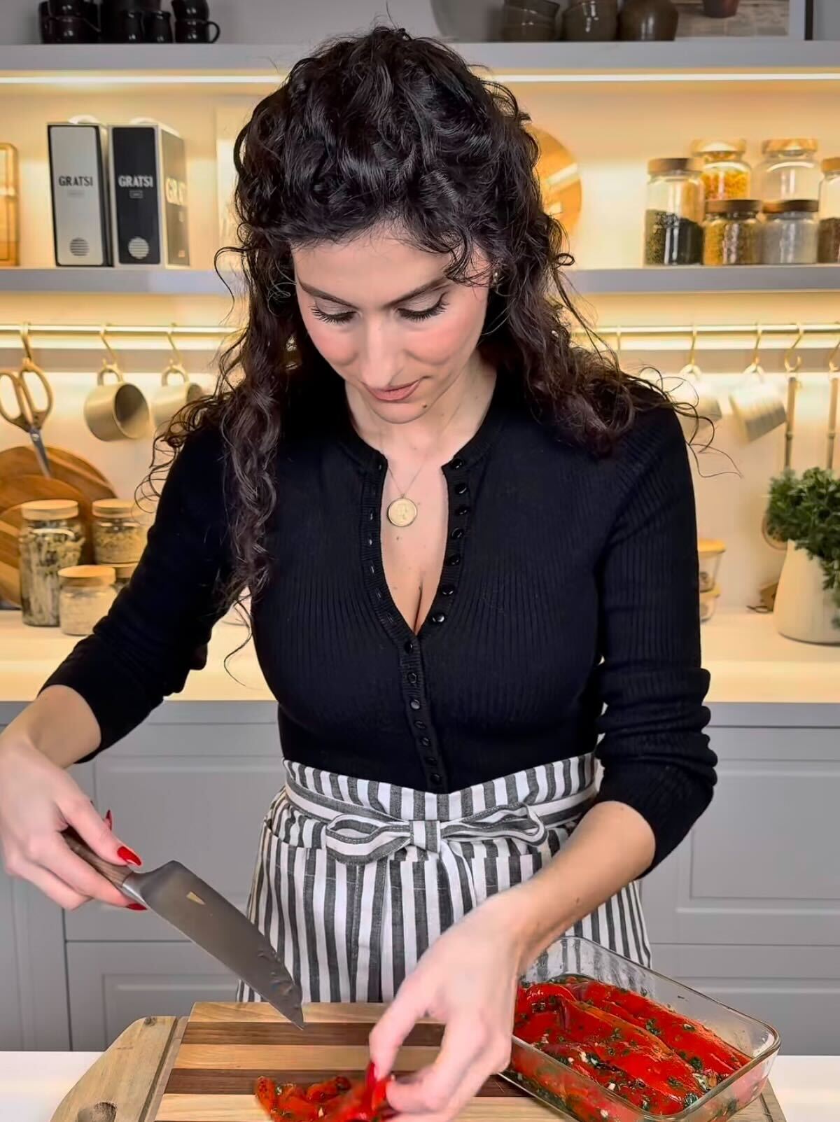 A woman with curly dark hair, wearing a black top and a striped apron, is slicing red peppers on a wooden cutting board in a modern kitchen with shelves holding jars, dishes, and utensils in the background.