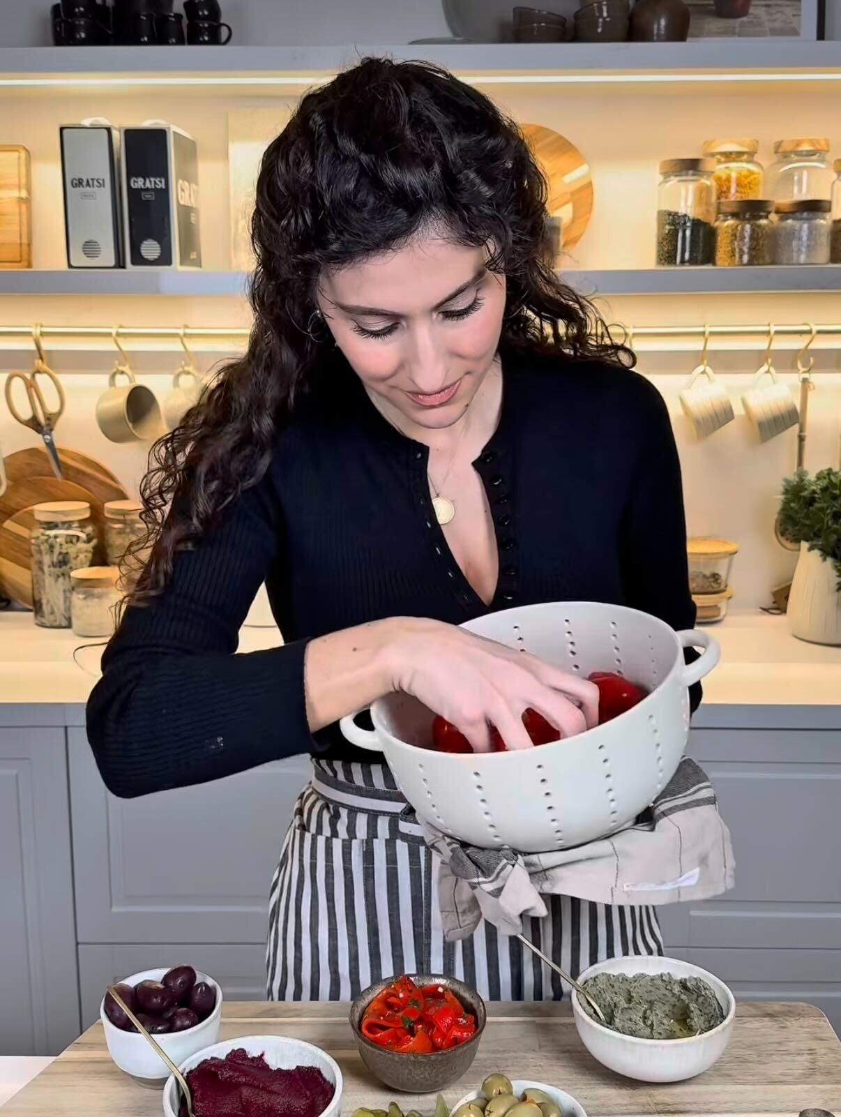 A woman with long curly hair stands in a kitchen, looking down as she places her hand into a white colander filled with red bell peppers. Various bowls of ingredients are arranged on the counter in front of her.