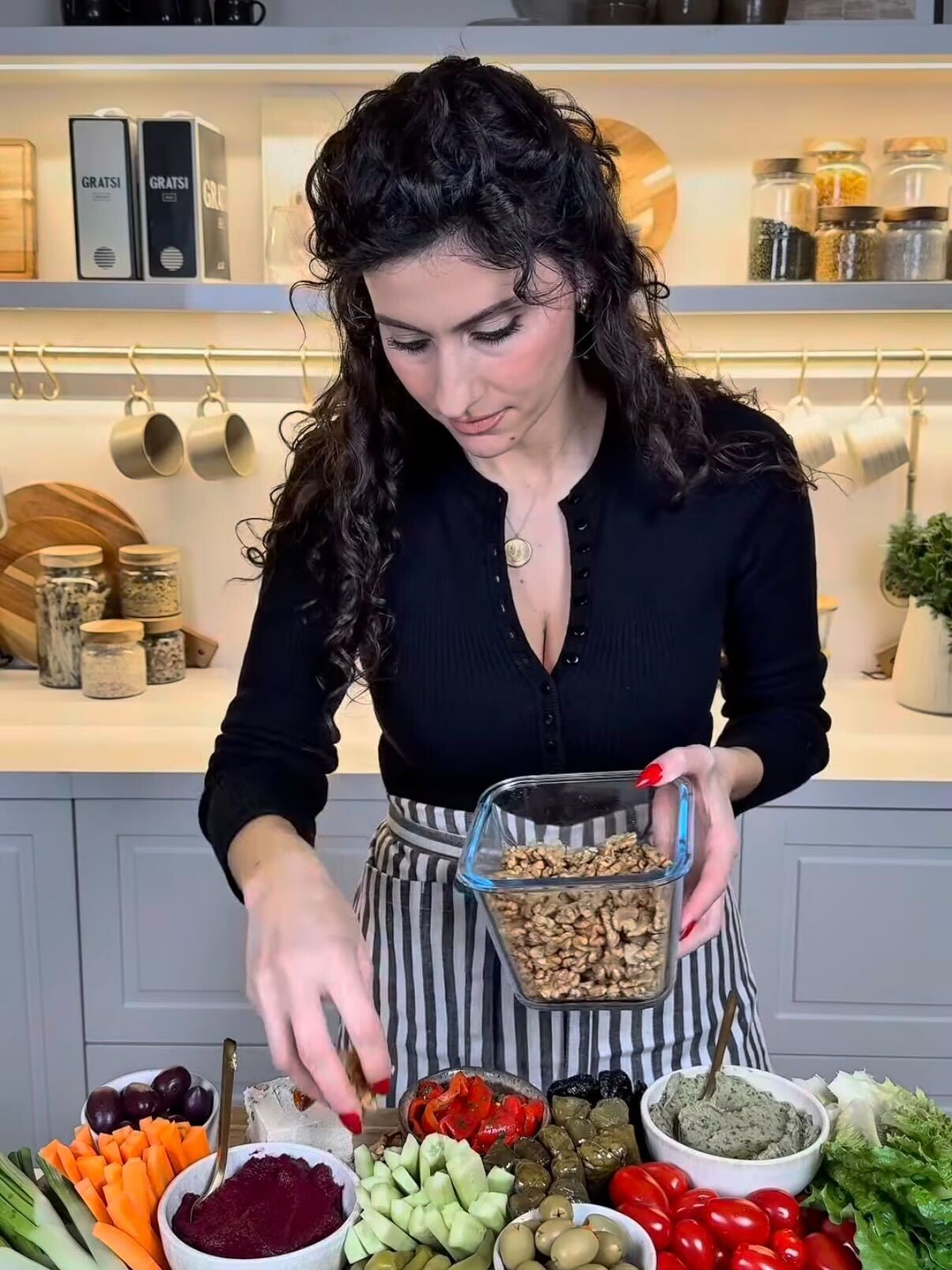 A woman with wavy dark hair prepares a vegetable platter in a kitchen, holding a container of walnuts and arranging assorted fresh veggies, dips, and olives on a counter in front of her.