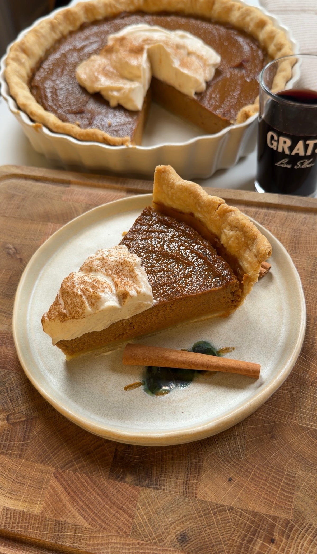A slice of pumpkin pie topped with whipped cream and cinnamon on a plate with a cinnamon stick, next to a whole pie and a glass of red wine.