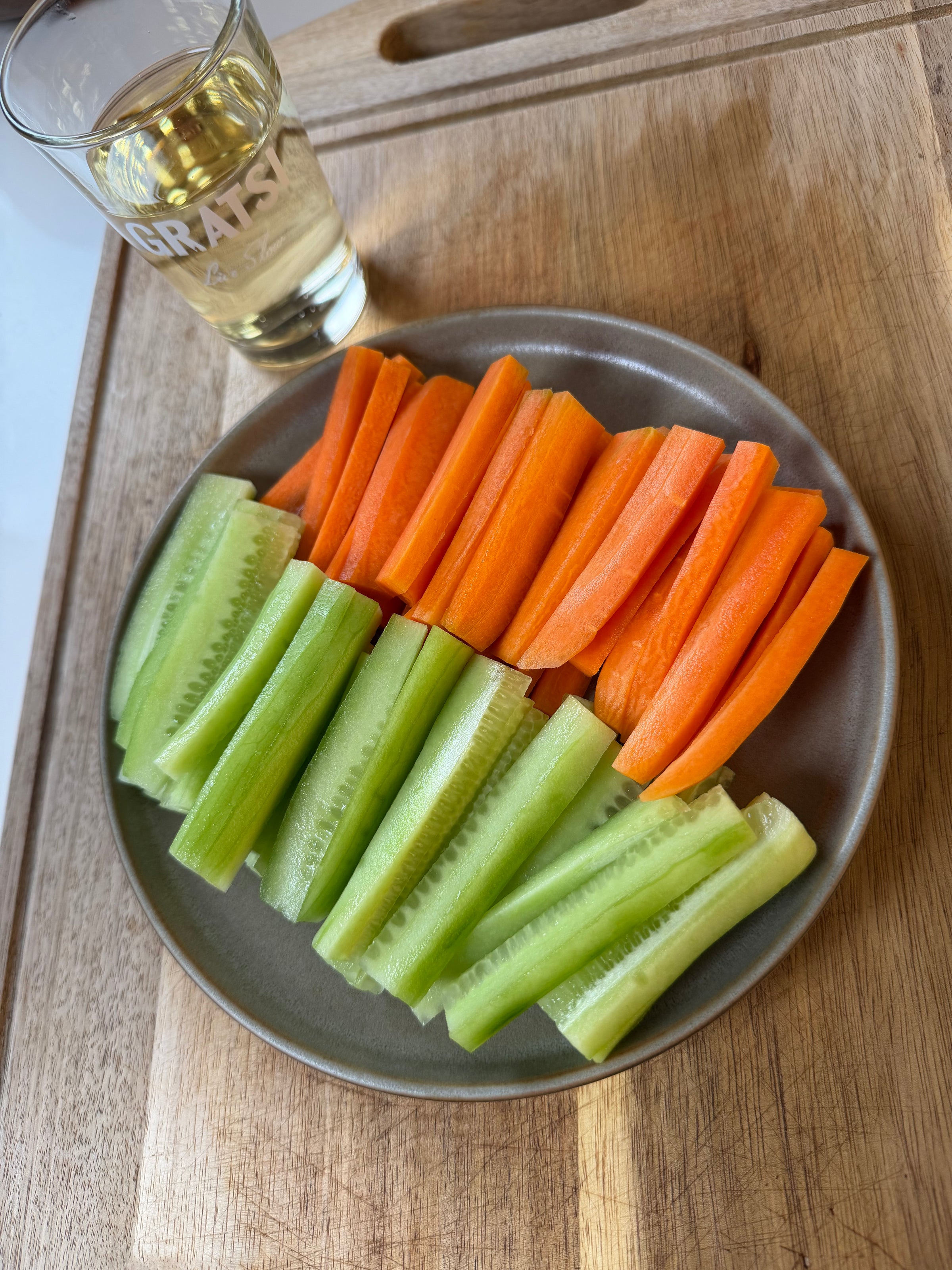 A plate with neatly arranged carrot, celery, and cucumber sticks sits on a wooden board. A glass of light-colored beverage is placed beside the plate.