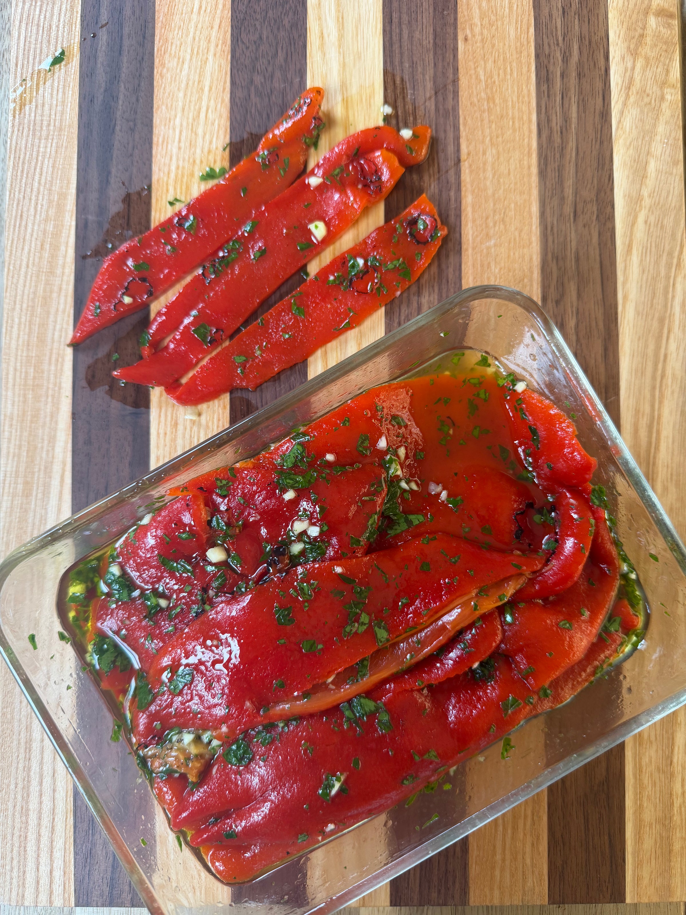 Sliced roasted red peppers marinated with herbs and garlic in a glass container, with additional pepper slices on a striped wooden cutting board.