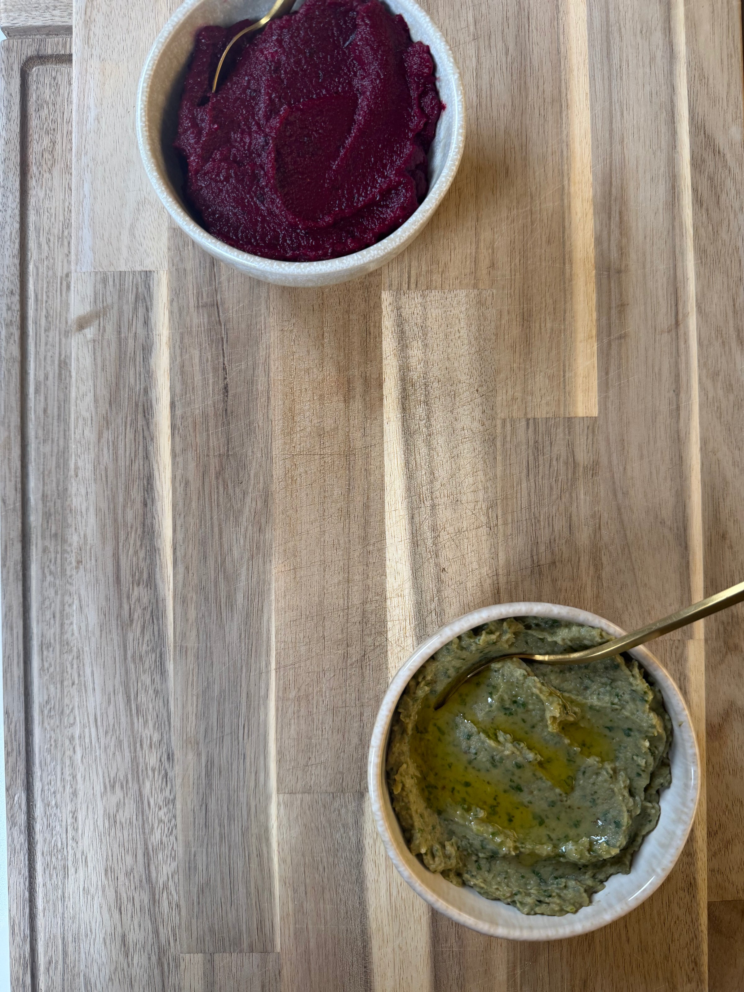 Two small bowls on a wooden surface: the top bowl contains a deep red spread (likely beetroot dip), and the bottom bowl holds a greenish dip drizzled with olive oil. Each bowl has a spoon inside.