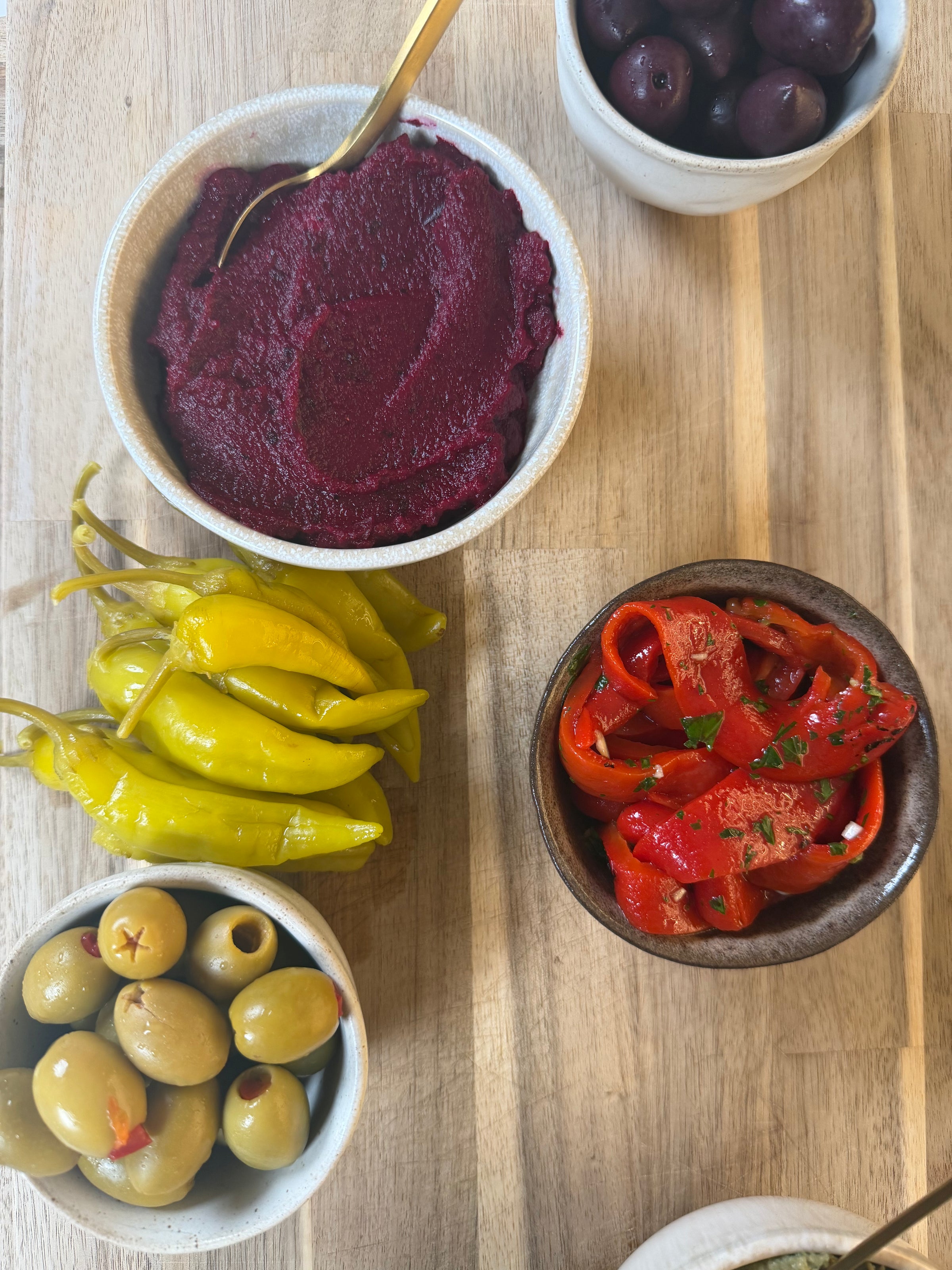 Small bowls on a wooden surface filled with various appetizers: green olives, yellow pickled peppers, sliced roasted red peppers with herbs, purple olives, and a bowl of deep red beet dip with a gold spoon.