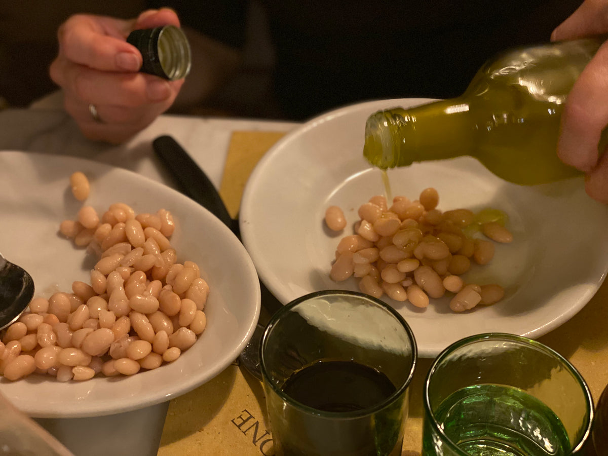 A person pours olive oil onto white beans on a plate, with another plate of beans, a spoon, and green glass cups visible on the table.