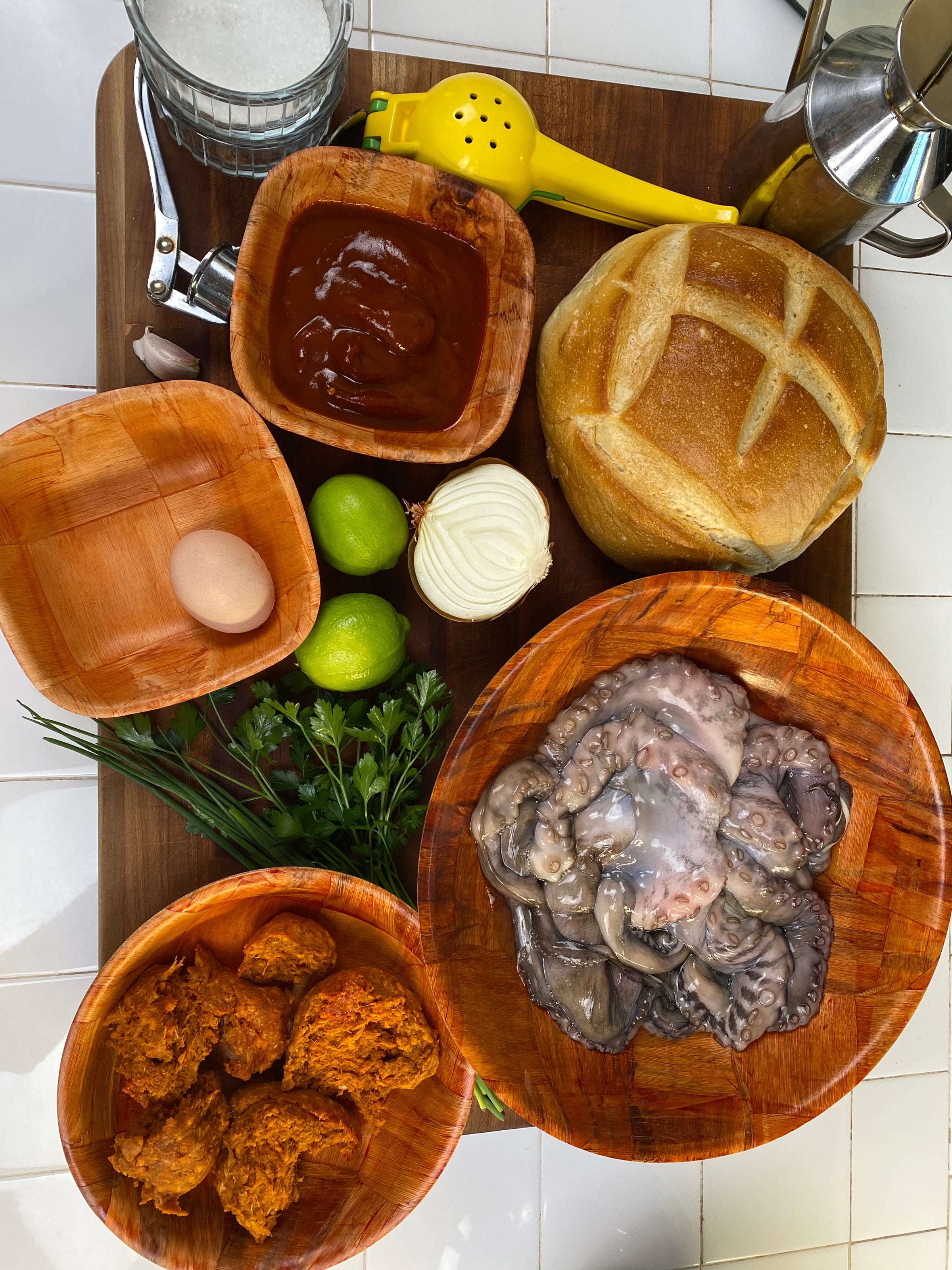 A wooden board displays octopus, a round loaf of bread, chorizo, an egg, limes, fresh herbs, a bowl of sauce, a sliced onion, a lemon squeezer, garlic, a metal oil bottle, and a glass of water.