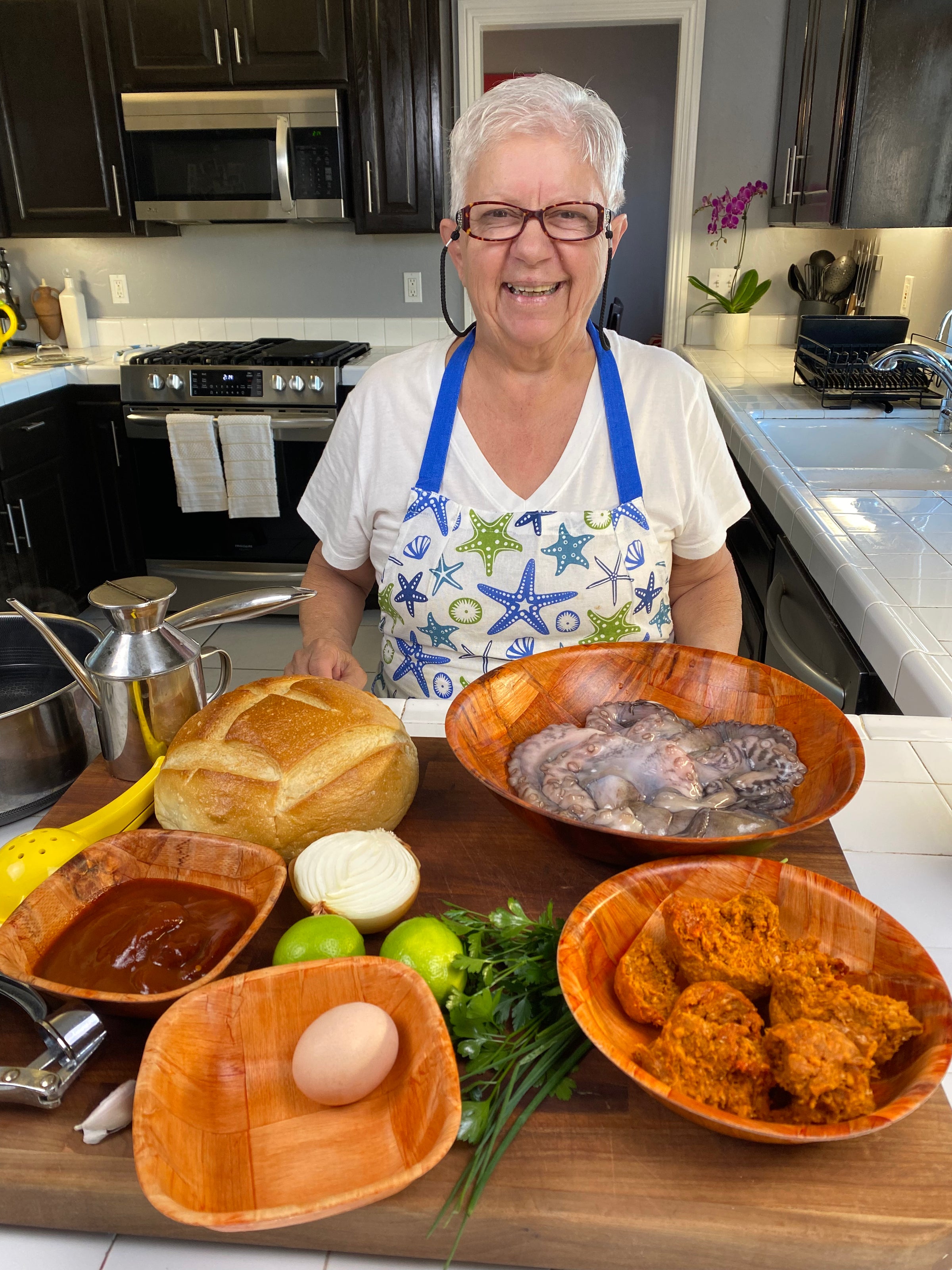 An older woman with short white hair, glasses, and a starfish-patterned apron smiles in a kitchen behind a counter with bread, octopus, onions, herbs, an egg, green tomatoes, and bowls of red sauce and chorizo.