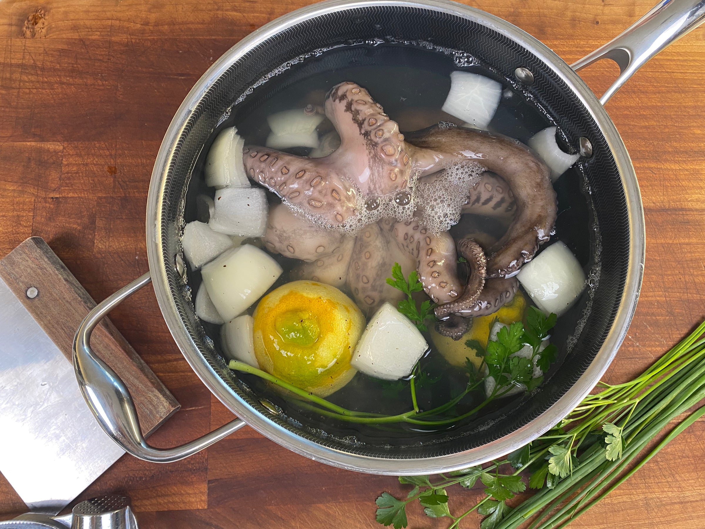 A pot on a wooden surface contains octopus tentacles, onion chunks, lemon halves, and fresh parsley submerged in water, ready for cooking. A cleaver and more parsley rest nearby.