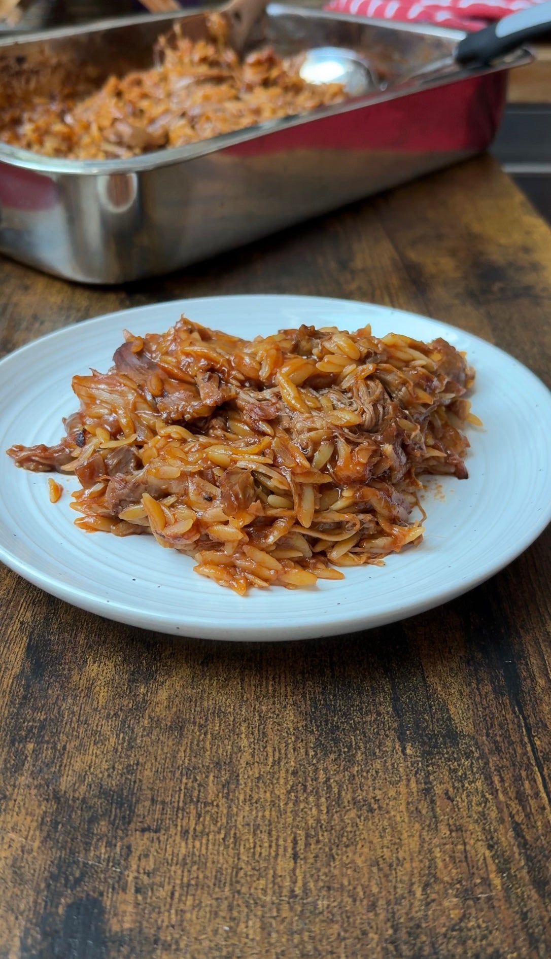 A white plate holds a serving of baked orzo pasta mixed with shredded meat and tomato sauce, sitting on a wooden table. A baking dish with more of the same dish is in the background.
