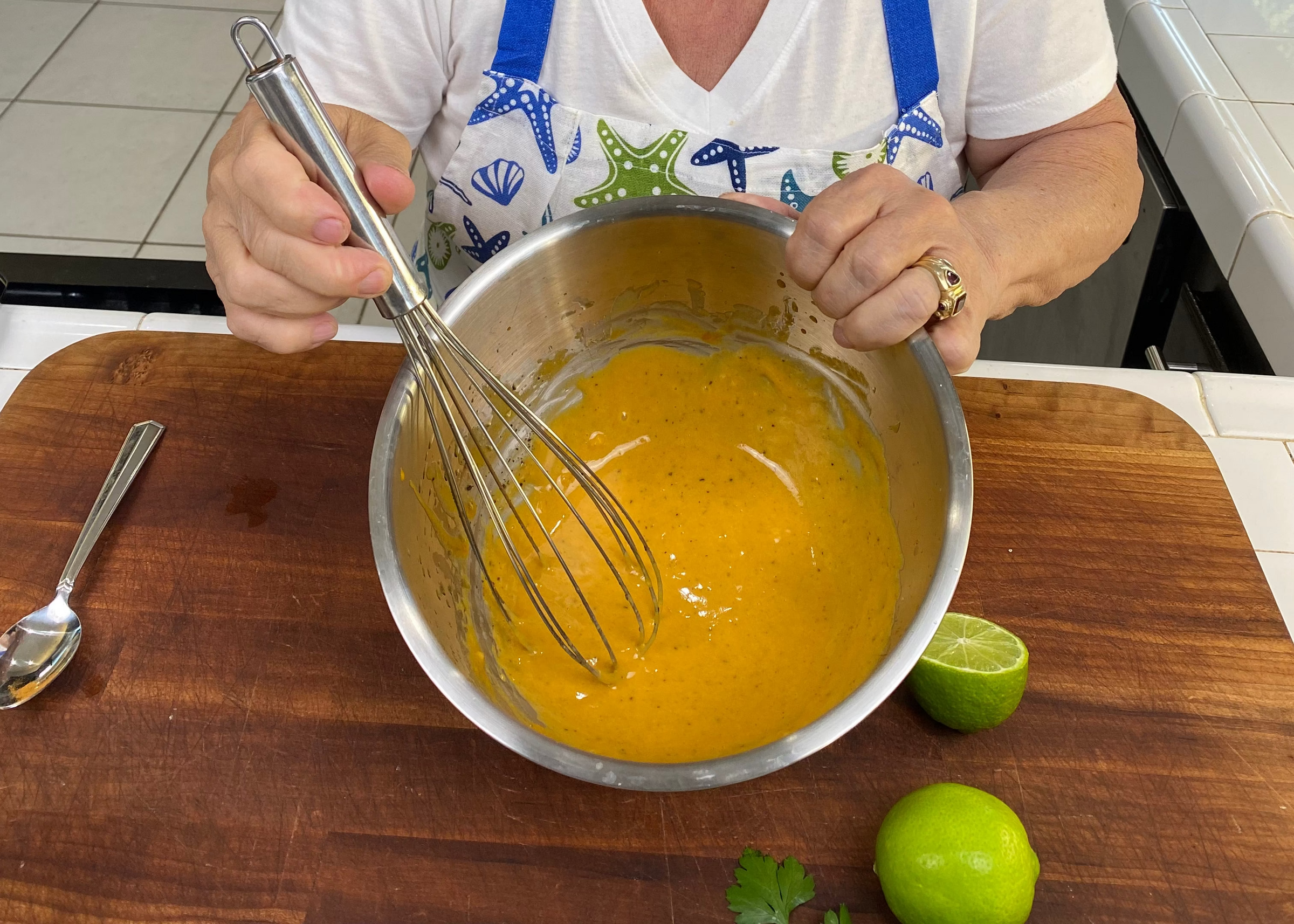 A person wearing a seashell-patterned apron is whisking a yellow mixture in a metal bowl on a wooden surface, with two limes—one cut in half—nearby.