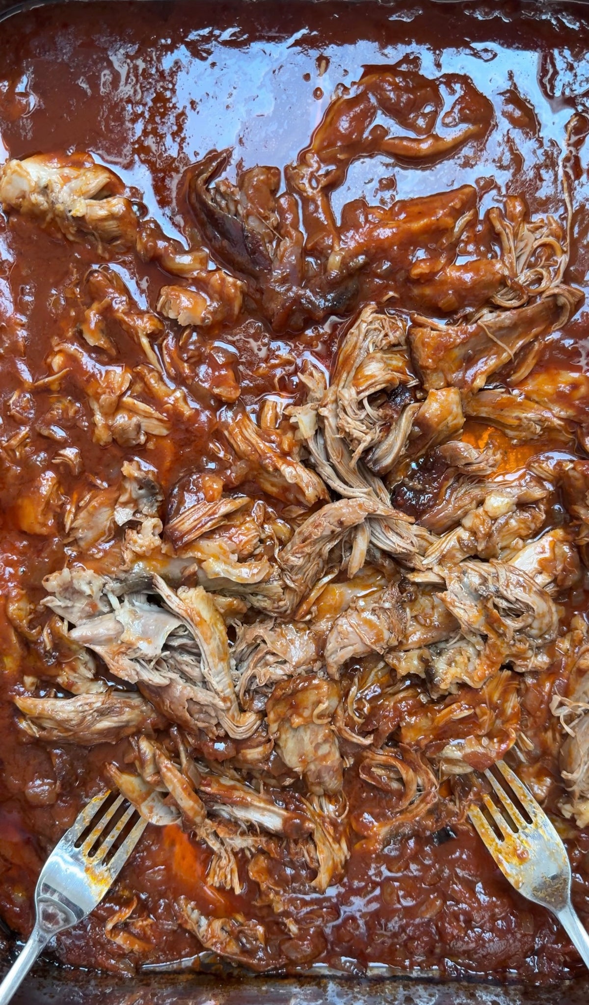 Two forks shred pieces of tender pulled pork in a pan filled with rich, reddish-brown barbecue sauce. The meat is juicy and coated in sauce, with strands separating easily.