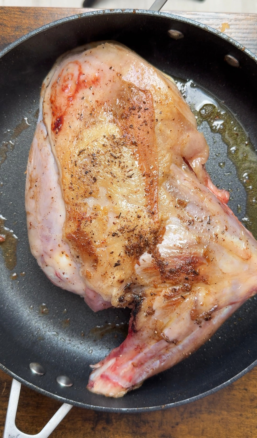 A large, seasoned piece of raw meat with some browning sits in a black frying pan with oil, ready to be cooked.