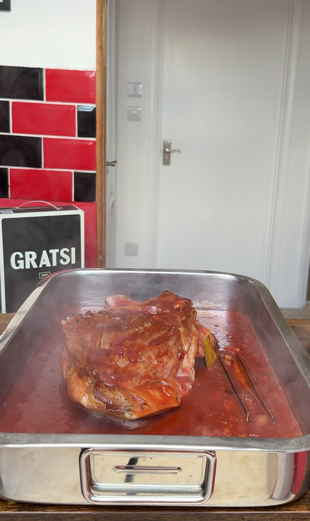 A roasted meat joint sits in a metal tray filled with tomato sauce and herbs, with steam rising. The background shows a kitchen with a GRATSI tin and red and black tiles.