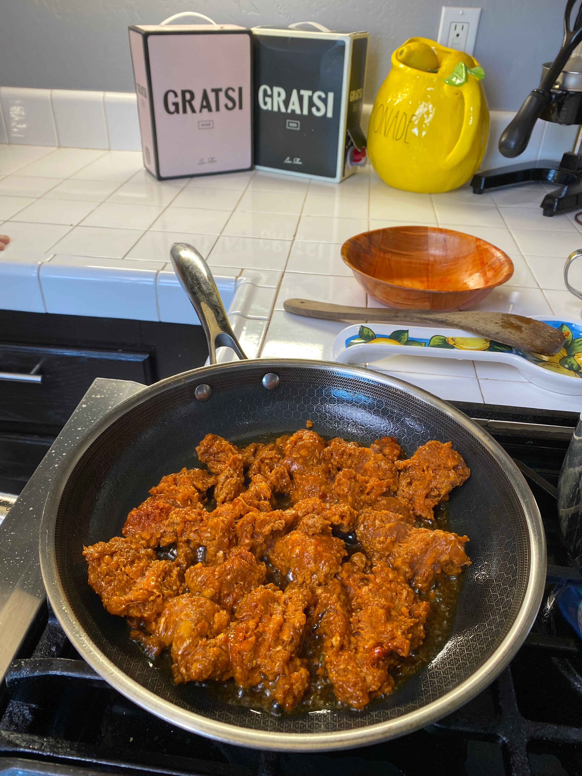 A frying pan on a stove contains sizzling, seasoned pieces of meat. On the white tiled counter behind, there are two Gratsi boxed wines, a yellow ceramic lemon container, and some kitchen utensils.