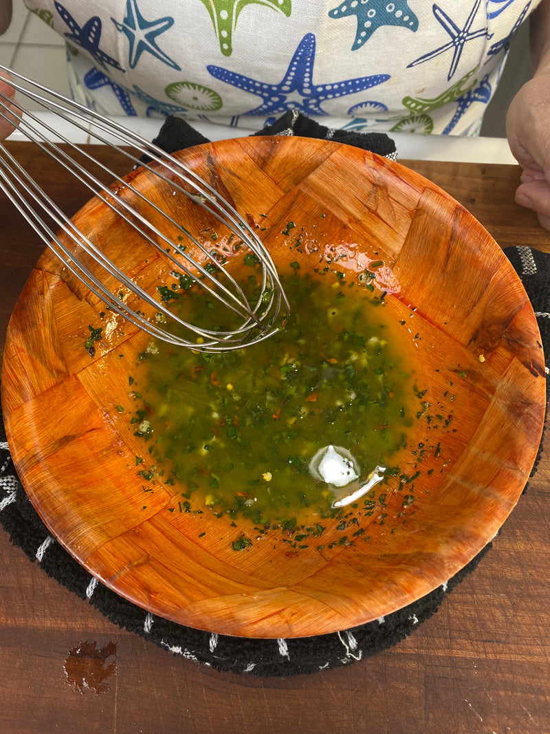 A person whisking a green herb vinaigrette in a wooden bowl, wearing a colorful starfish-patterned apron. The bowl is on a dark towel and wooden surface.