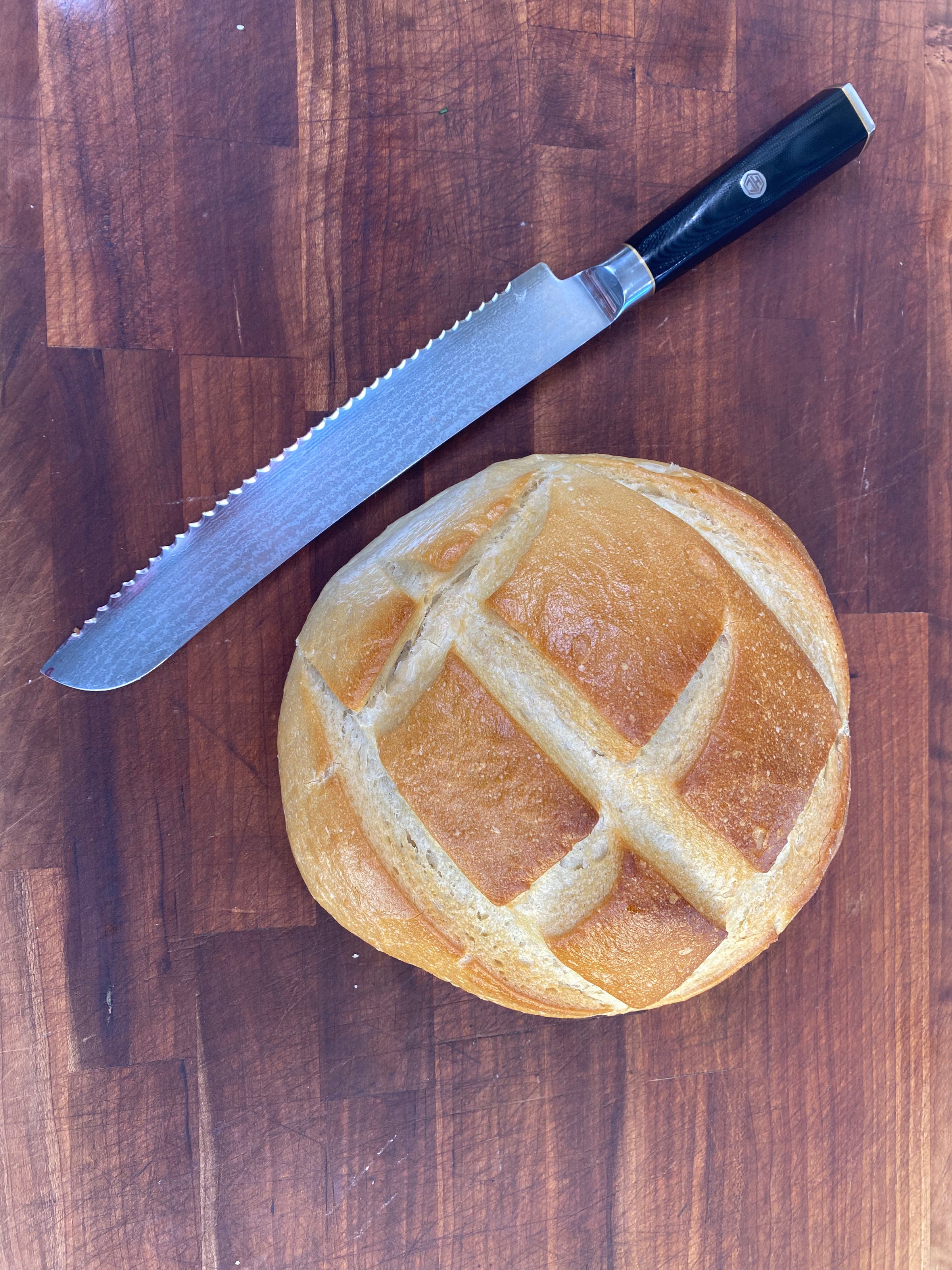 A round loaf of bread with a golden crust and an X scored on top sits on a wooden cutting board next to a serrated bread knife with a black handle.