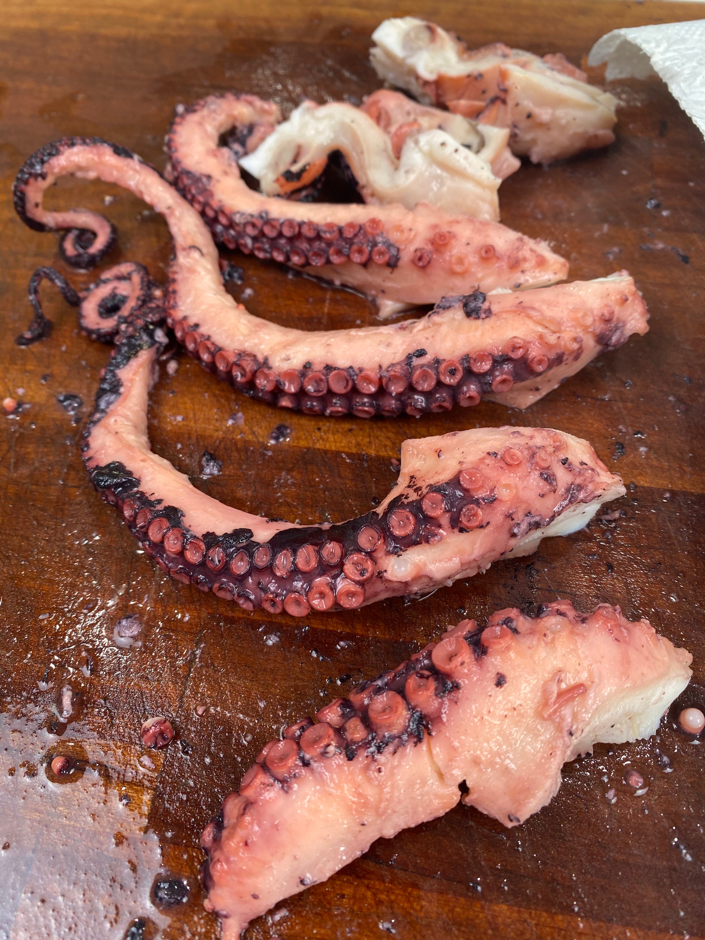 Several cooked octopus tentacles are laid out on a wooden cutting board. The tentacles have a pinkish color with suction cups visible and some juices on the board.