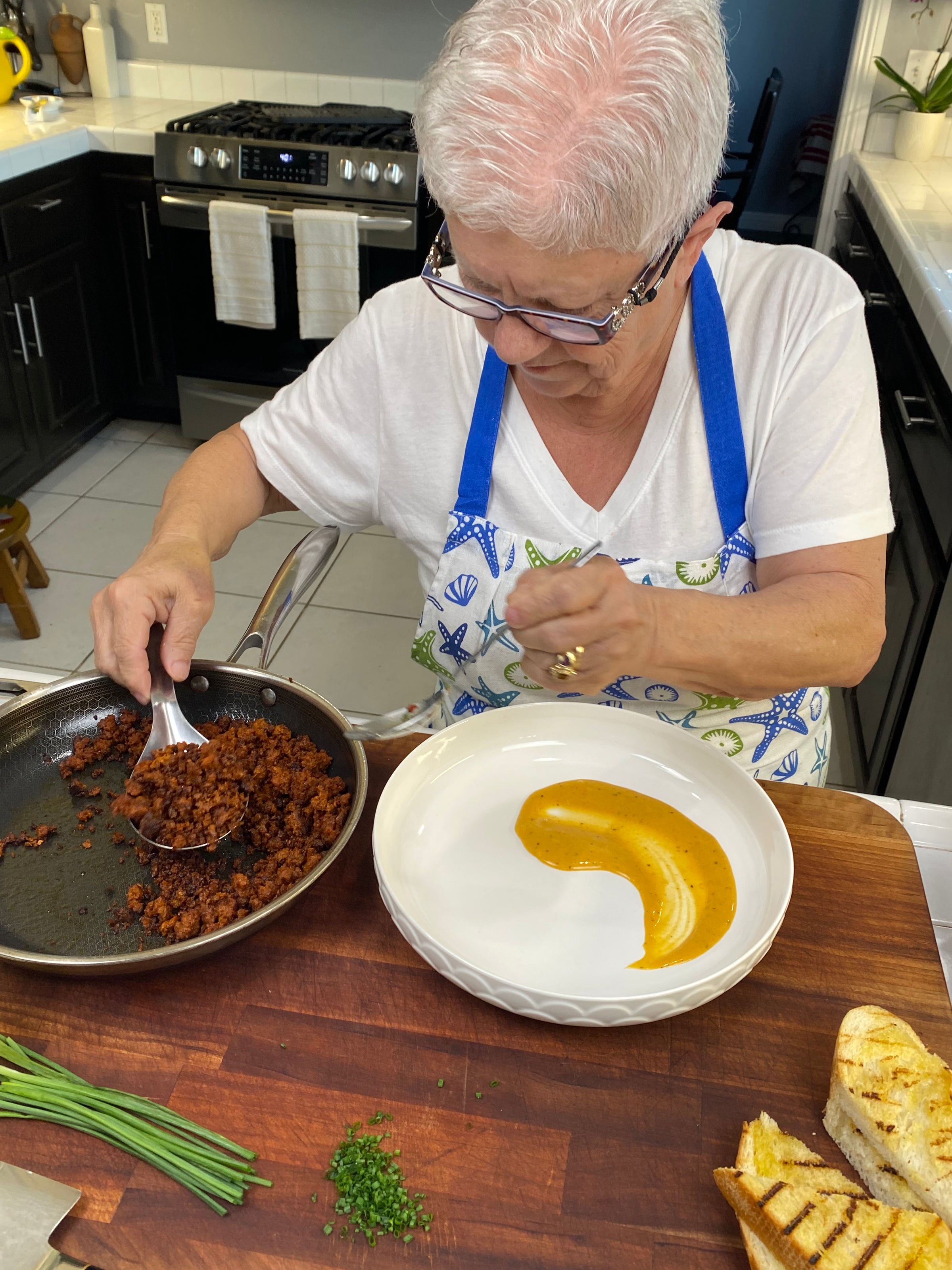 An elderly woman with short white hair prepares a dish in a kitchen, spooning cooked ground meat from a pan onto a white plate with a swirl of yellow sauce. Chopped herbs and grilled bread are on the wooden counter nearby.