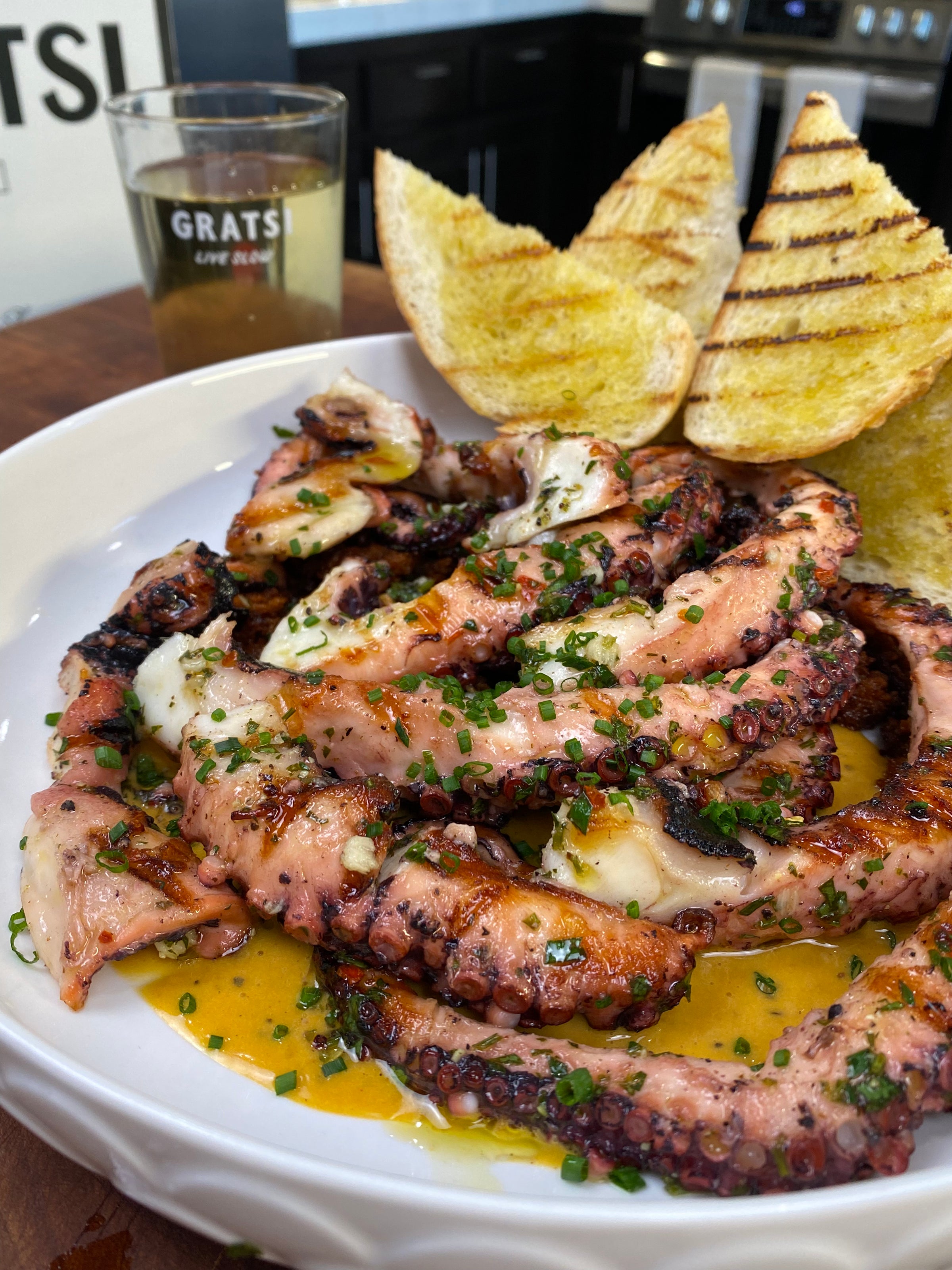 Grilled octopus tentacles topped with chopped chives, served with grilled bread slices and a glass of white wine on a wooden table. The dish is drizzled with a golden sauce.