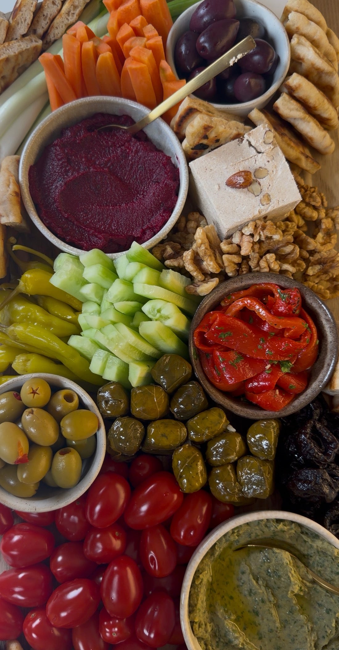 A colorful mezze platter with cucumber slices, cherry tomatoes, olives, grape leaves, red pepper strips, walnuts, hummus, beet dip, carrot sticks, pita bread, and assorted crackers.