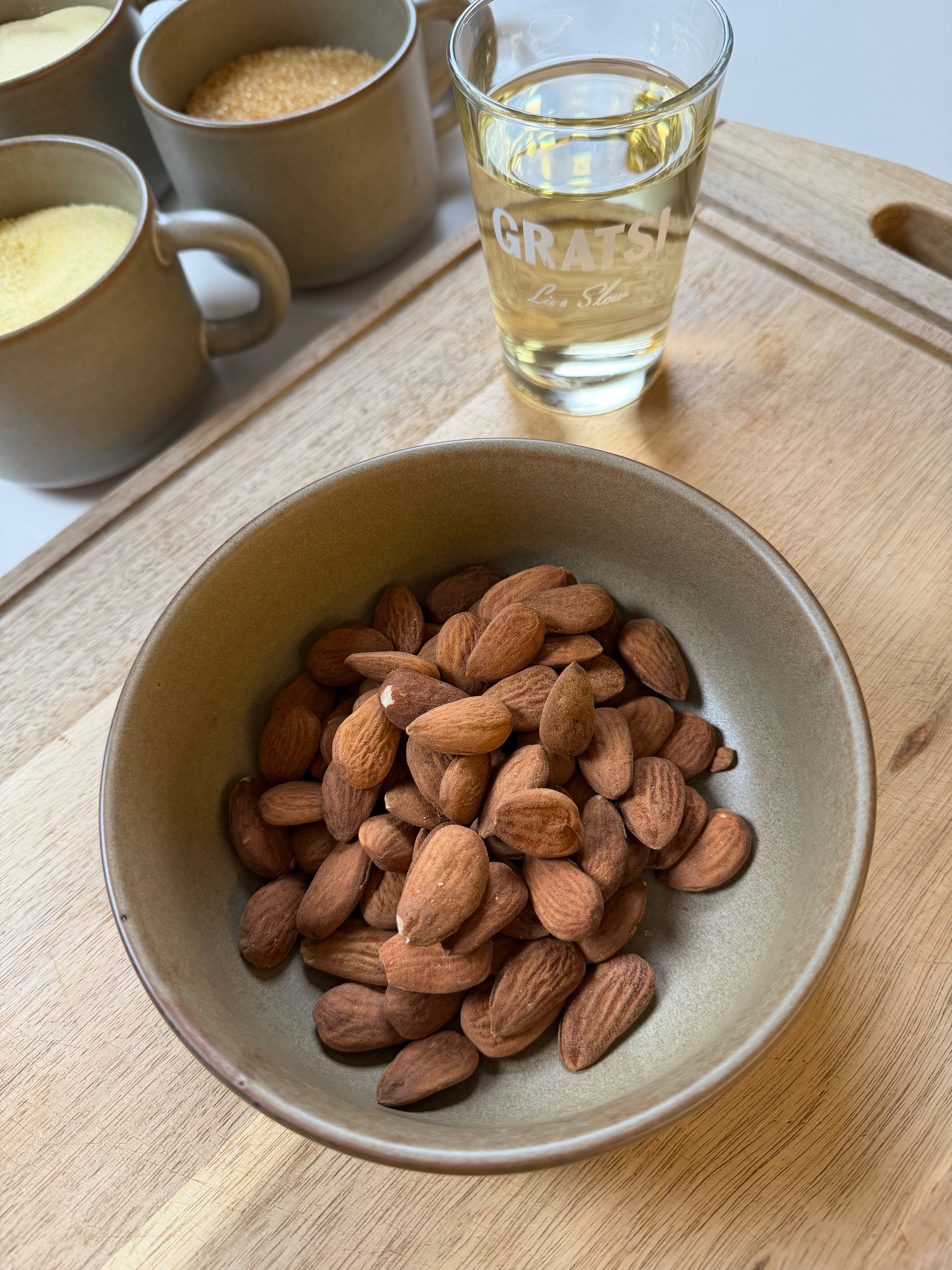 A bowl of whole almonds sits on a wooden surface next to a glass of clear liquid and three mugs filled with different granular ingredients.