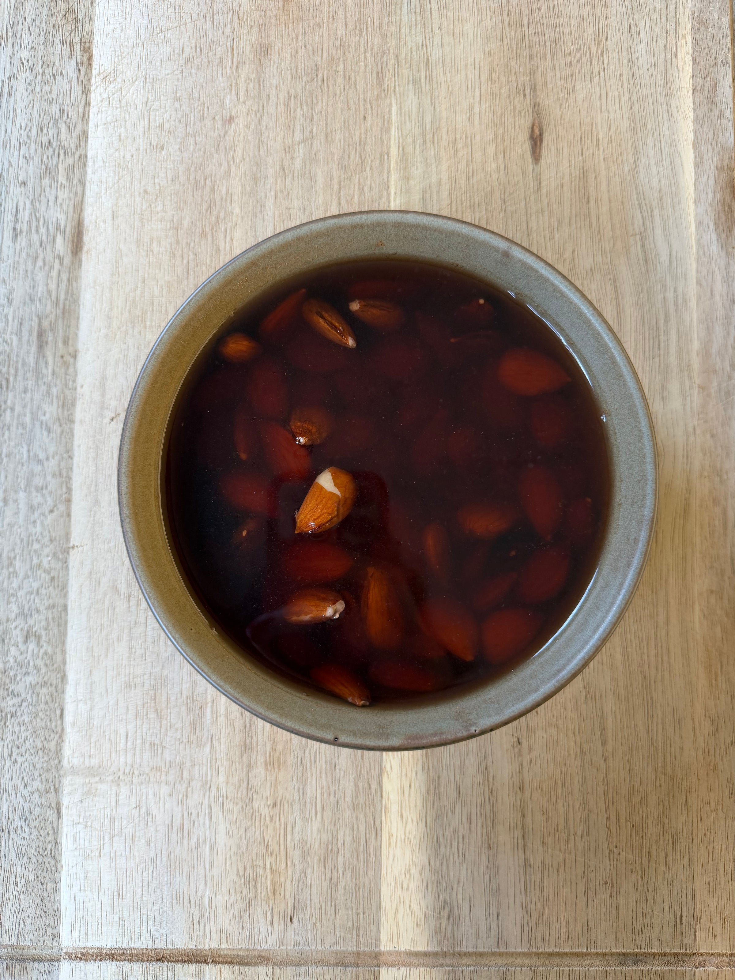 A brown bowl filled with dark liquid and whole almonds, placed on a light wooden surface.