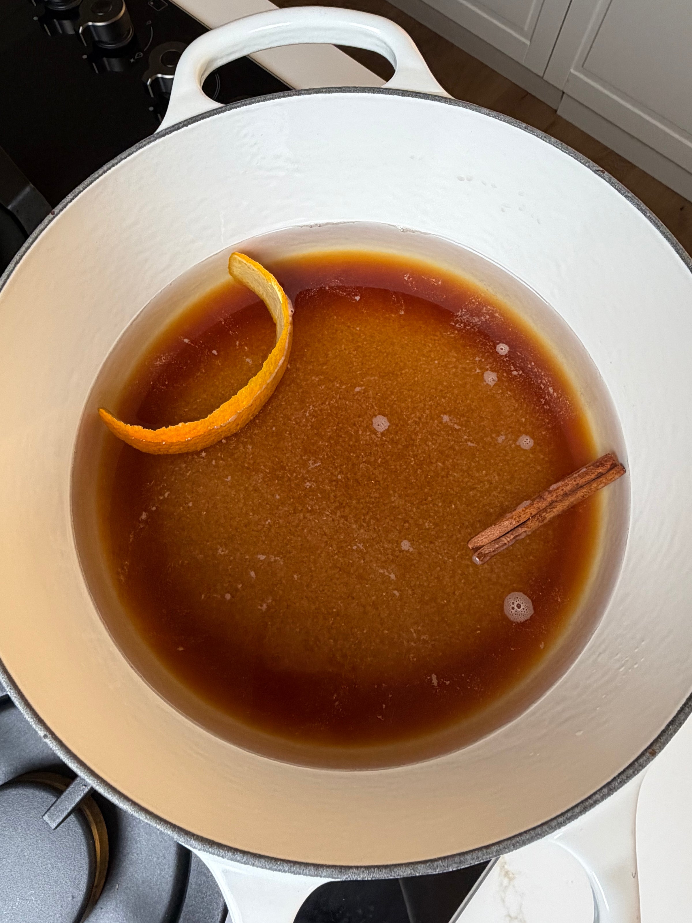 A white pot on a stovetop contains a brown liquid with a cinnamon stick and an orange peel floating in it.