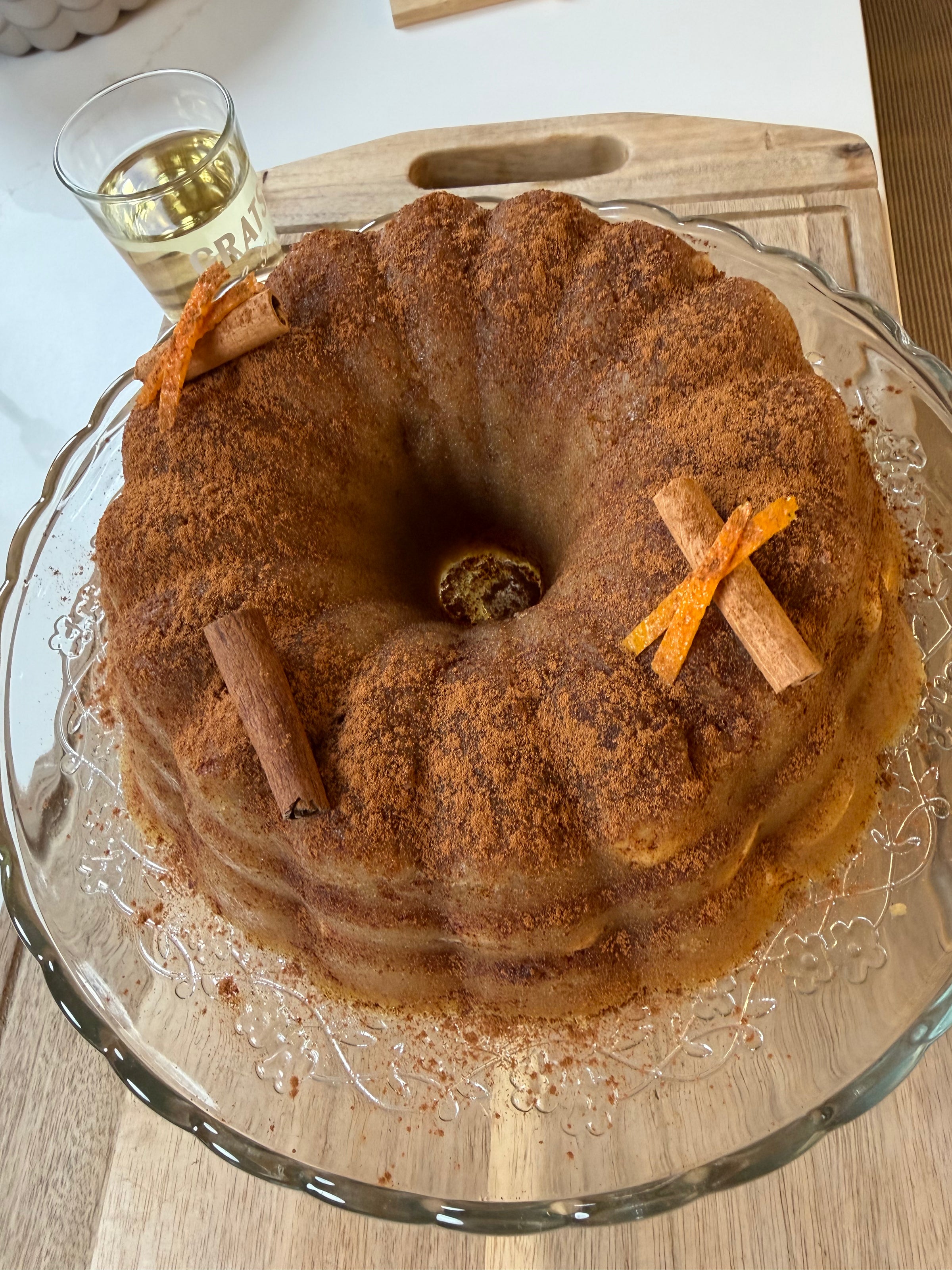 A round bundt cake dusted with cinnamon sits on a glass platter. It is decorated with cinnamon sticks and orange peel strips. In the background, there is a glass of liquid on a wooden surface.