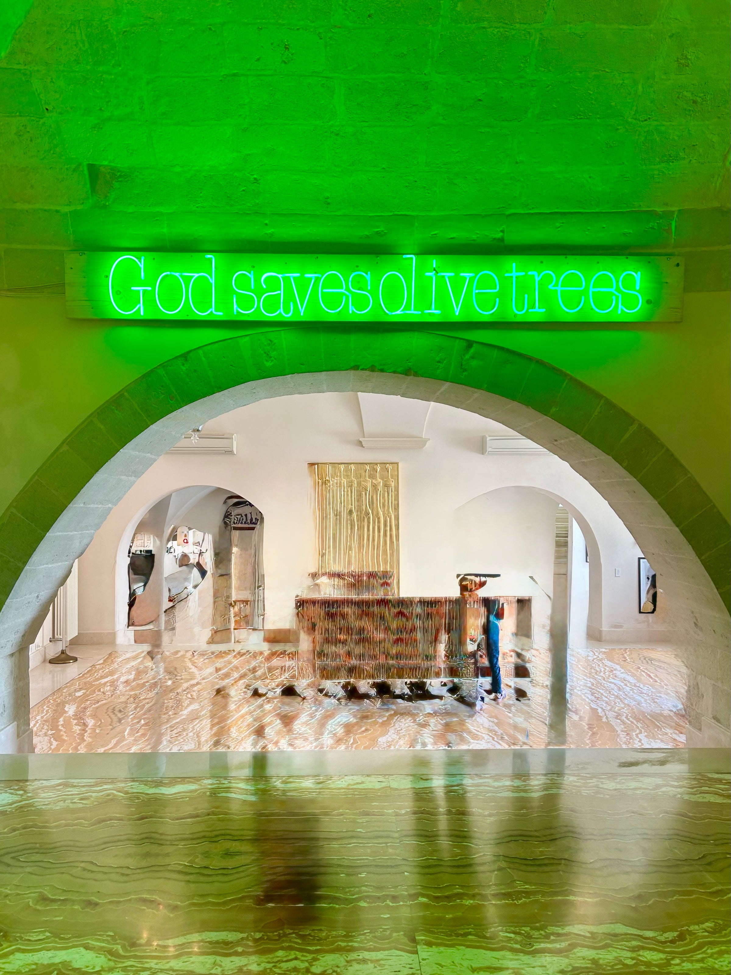 A neon green sign reading God saves olive trees hangs above a stone archway in a modern, art-filled room with marble-patterned floors and contemporary decor.