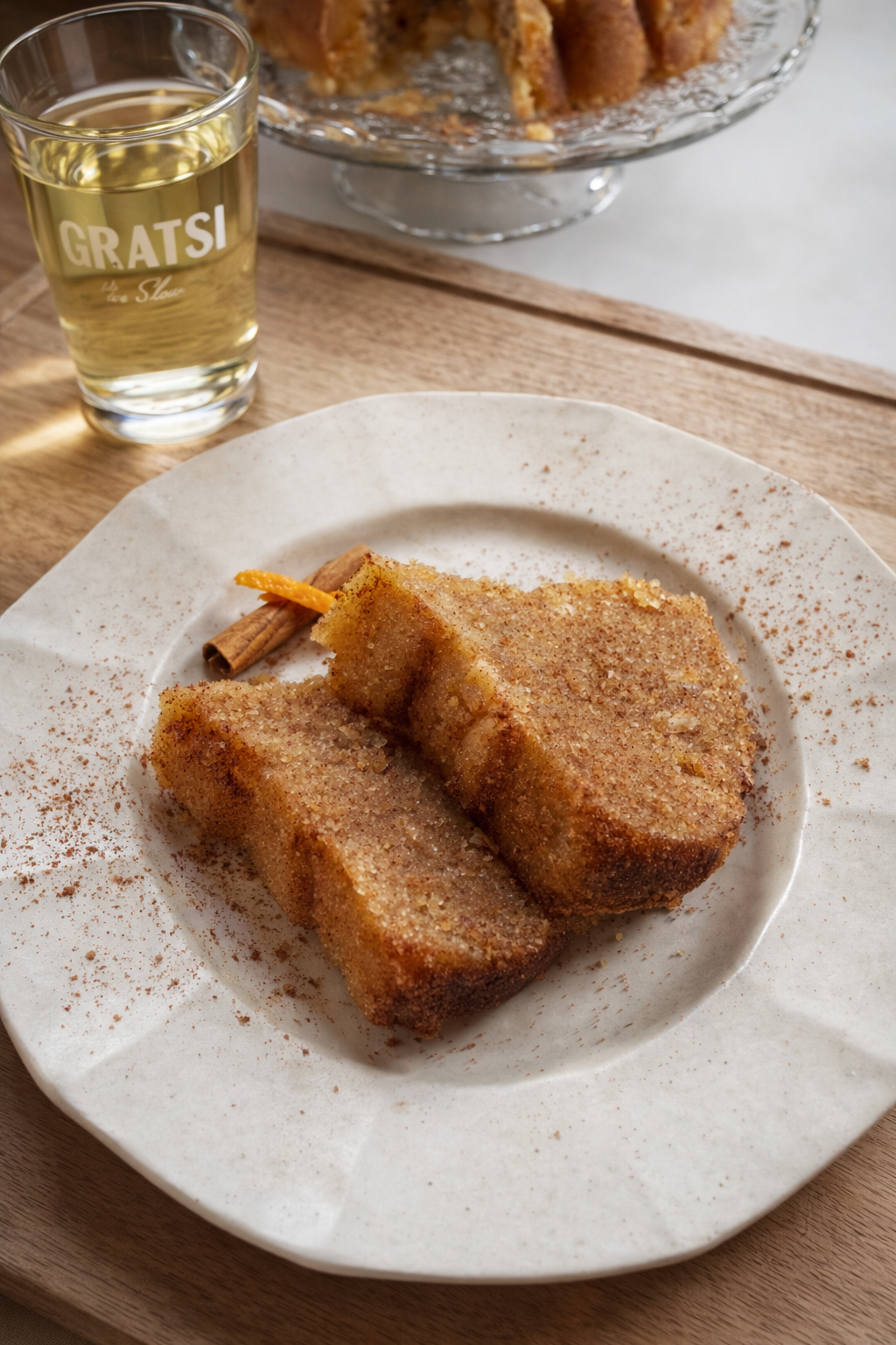 Two slices of cinnamon sugar cake on a white plate with a cinnamon stick, next to a glass of light-colored beverage on a wooden tray. A whole cake is visible in the background.