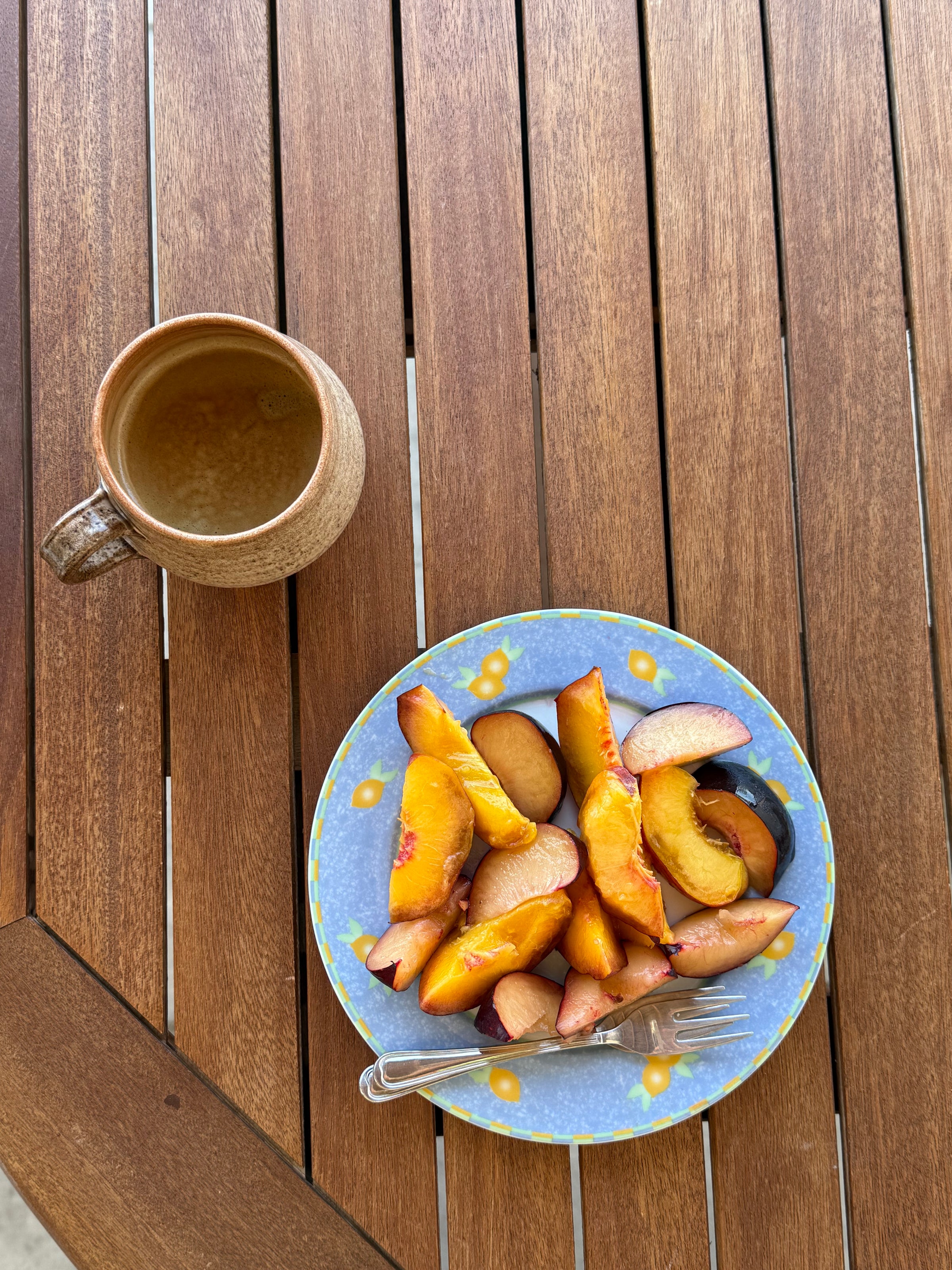 A brown mug next to a blue plate with sliced peaches and a fork sits on a wooden table.