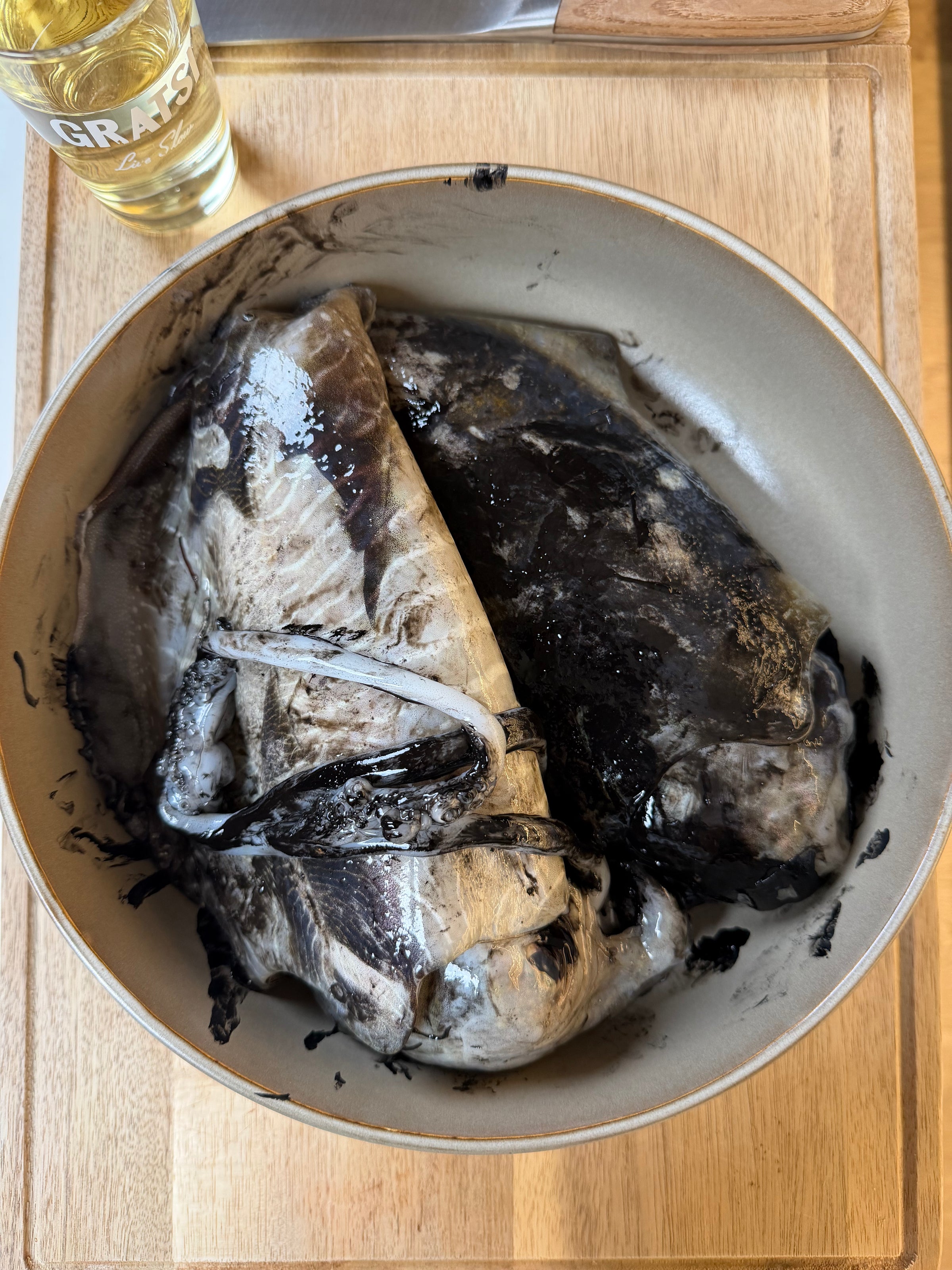 Two whole fish covered in black ink are placed in a beige bowl on a wooden surface, with part of a glass of white liquid visible at the top left corner.