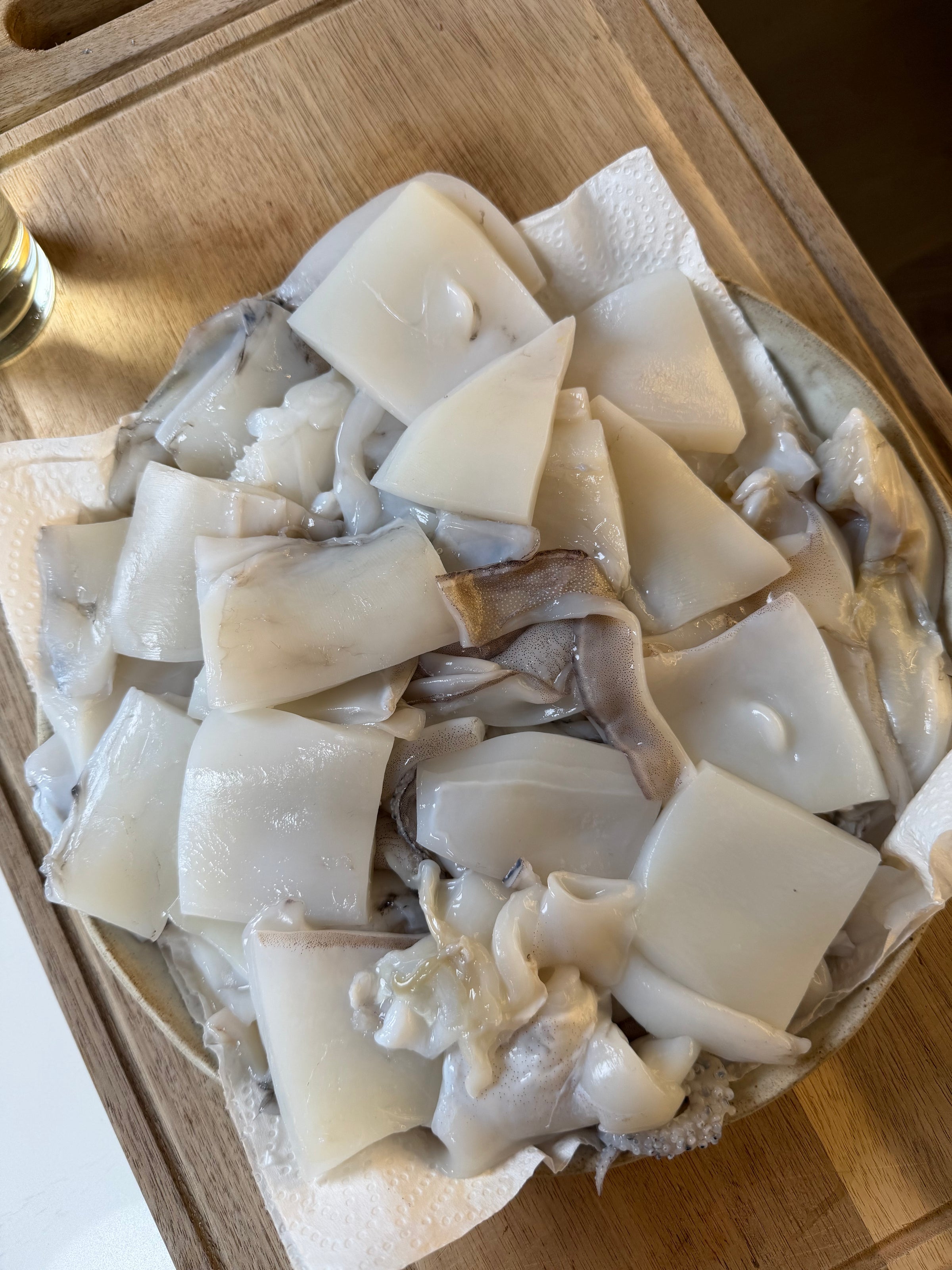 Freshly cut pieces of raw squid are arranged on a plate lined with paper towels, placed on a wooden cutting board under natural light.