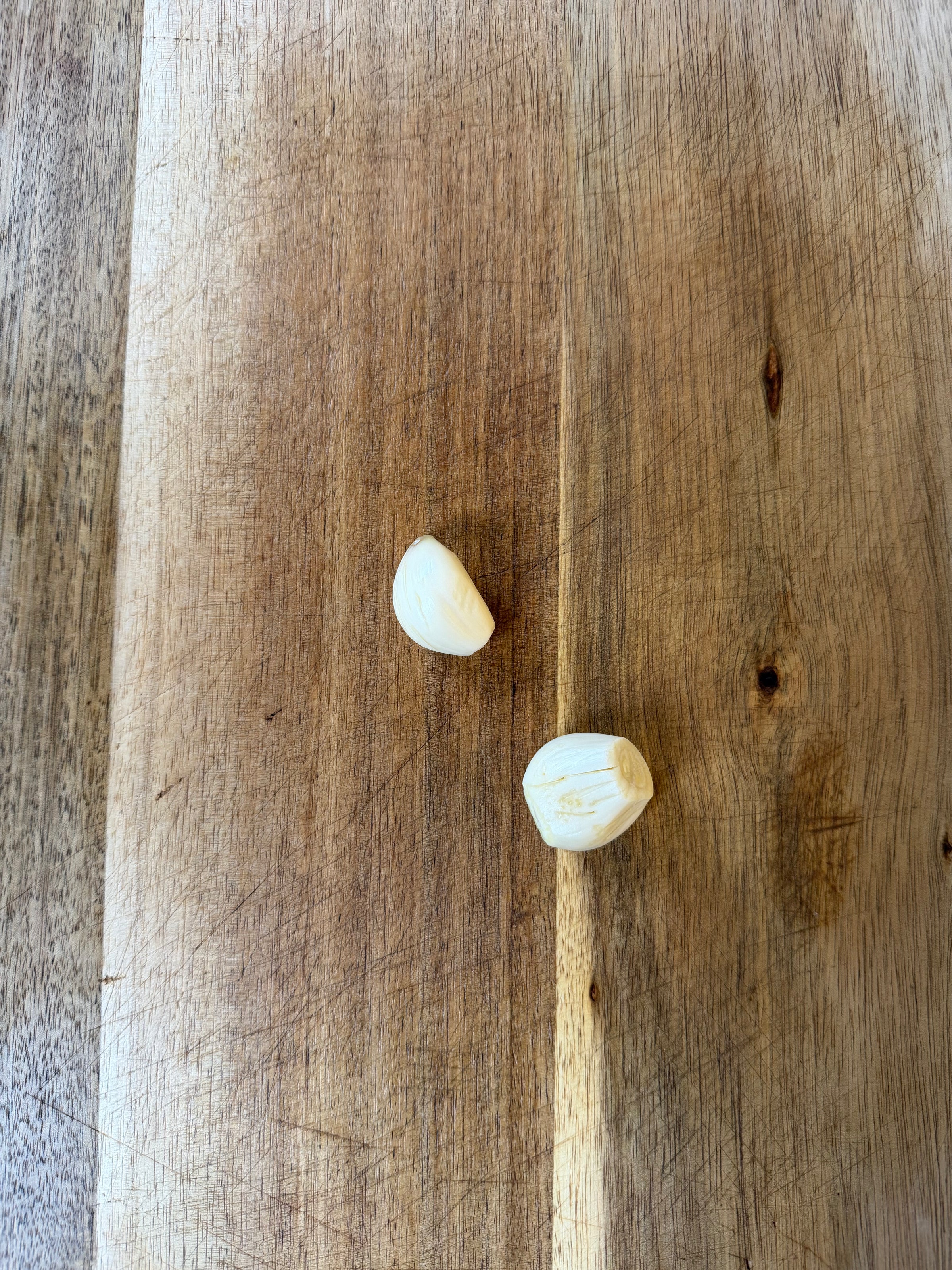 Two peeled garlic cloves sitting on a wooden cutting board with visible grain patterns.