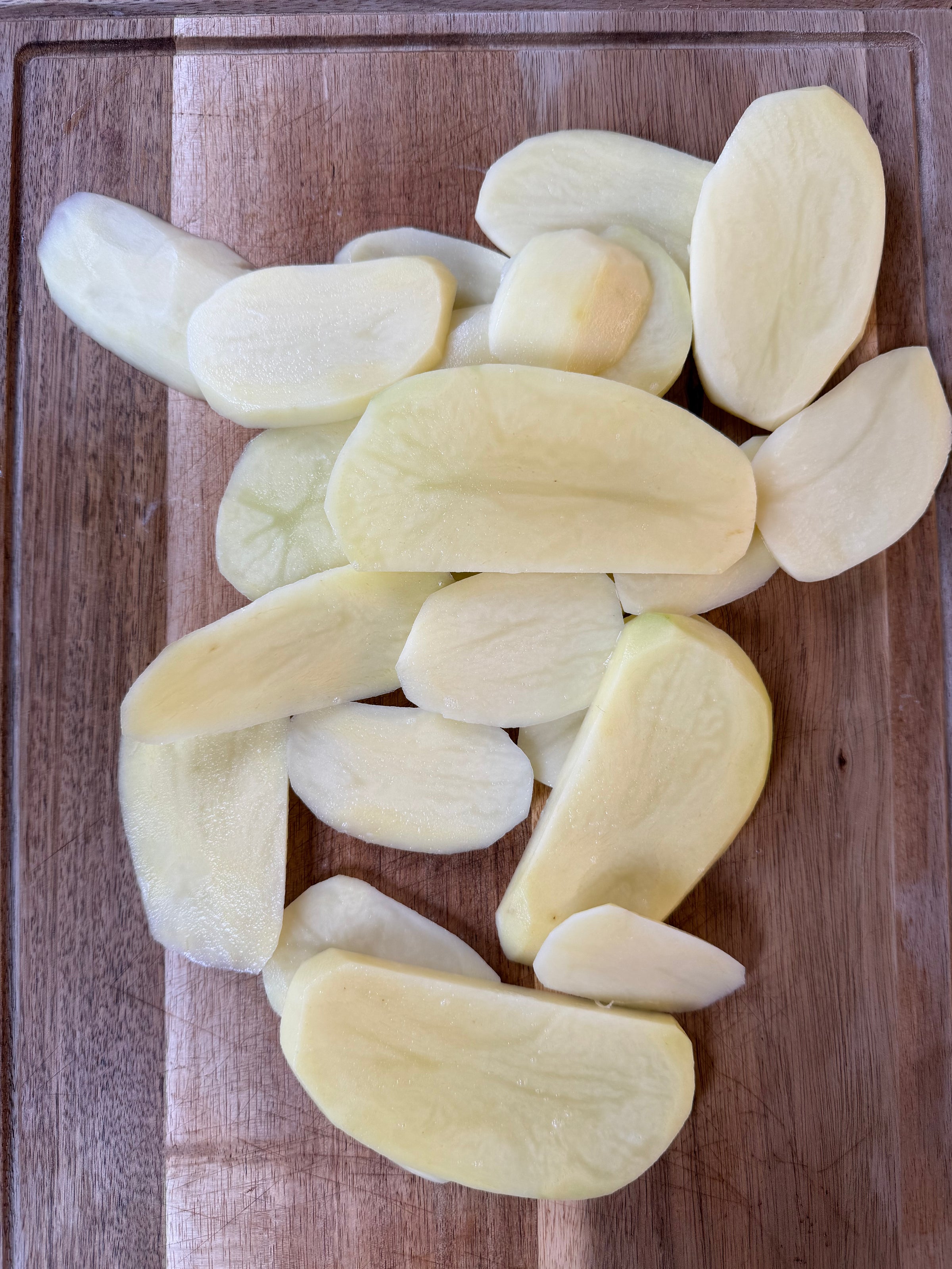 Thin slices of raw potato are spread out on a wooden cutting board, ready for cooking or further preparation.