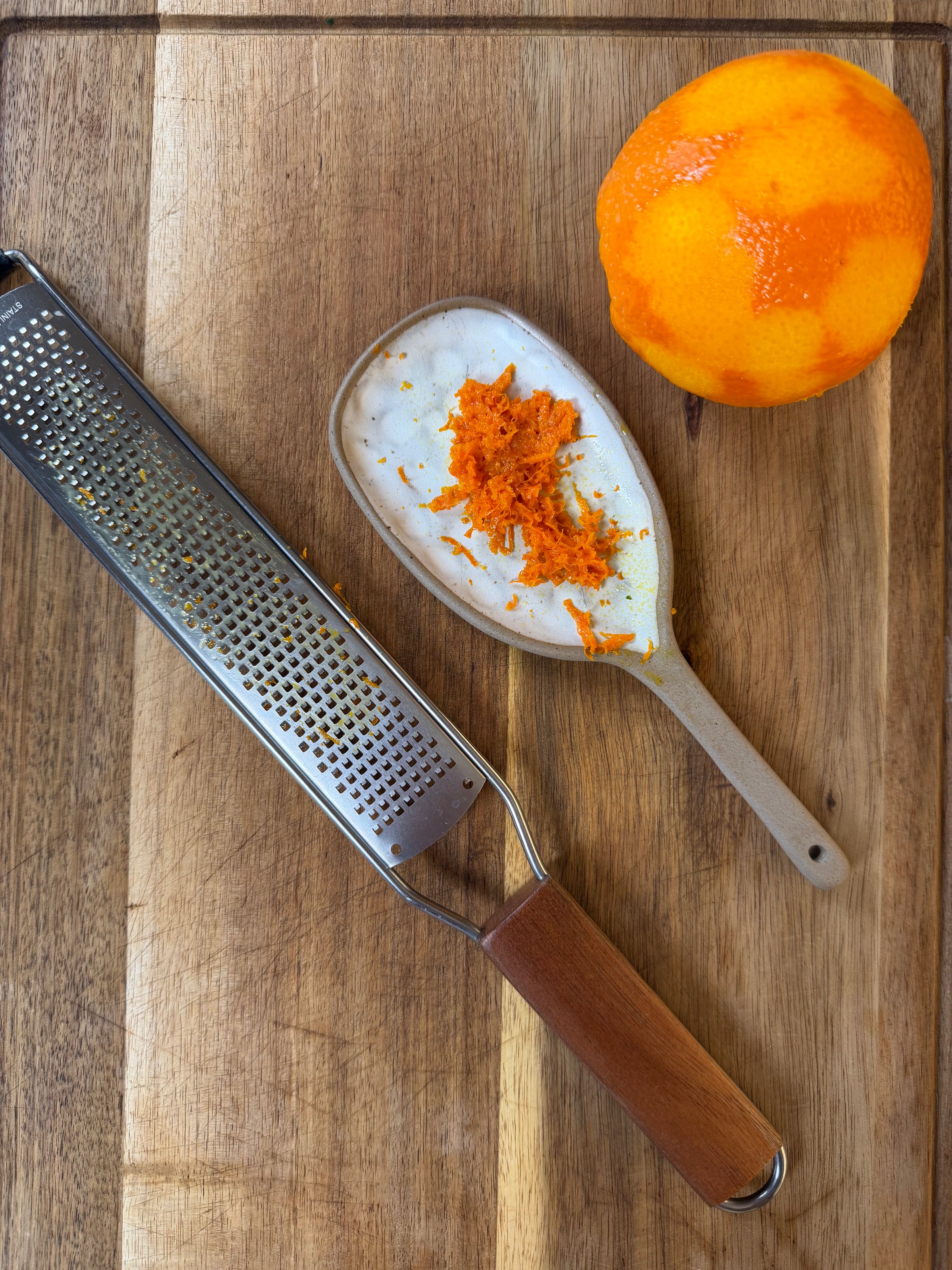 A microplane grater, a ceramic spoon with orange zest, and a partially zested orange sit on a wooden cutting board.
