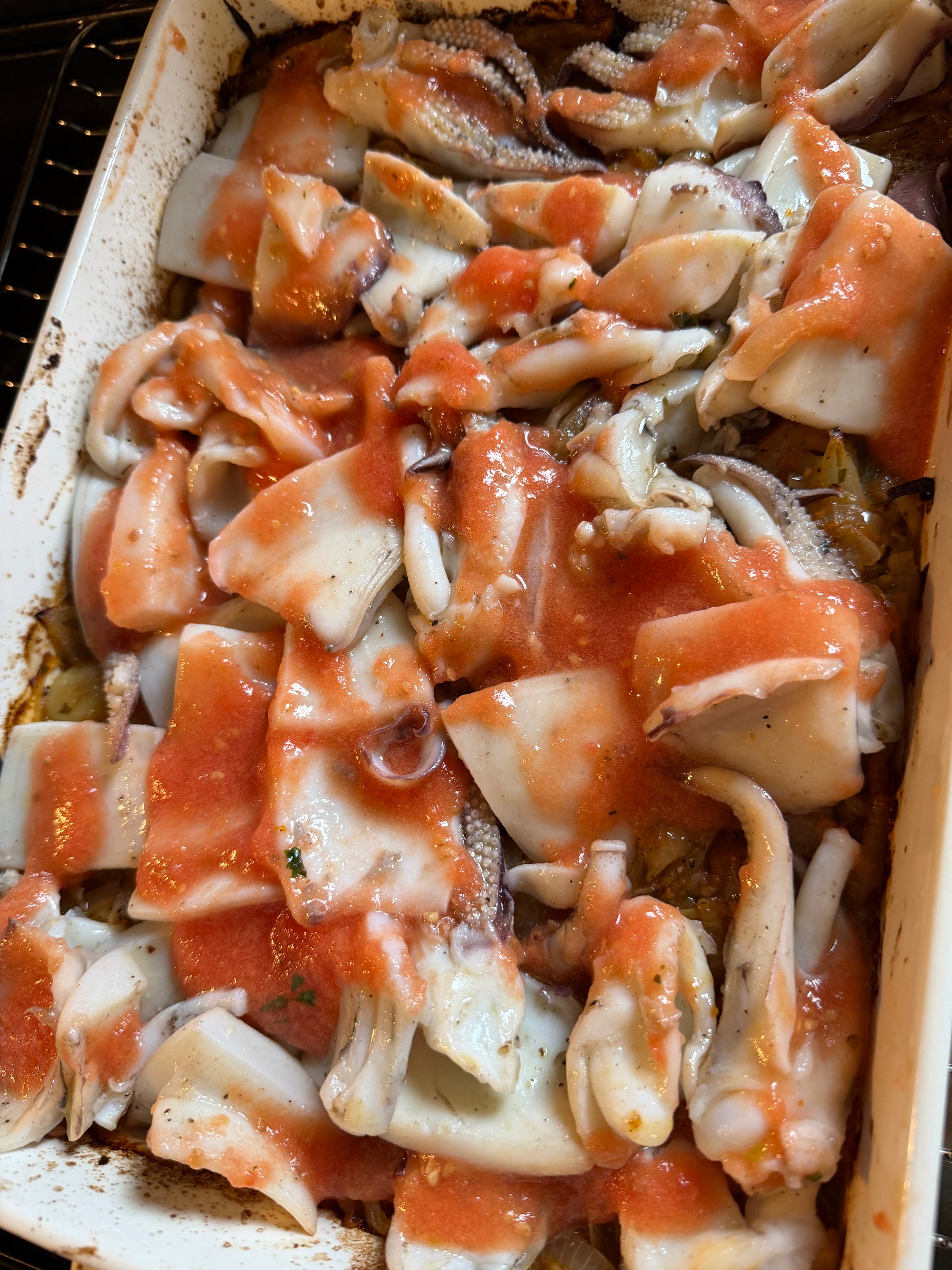Raw pieces of squid are spread in a baking dish and covered with a layer of tomato sauce, ready to be cooked in the oven.